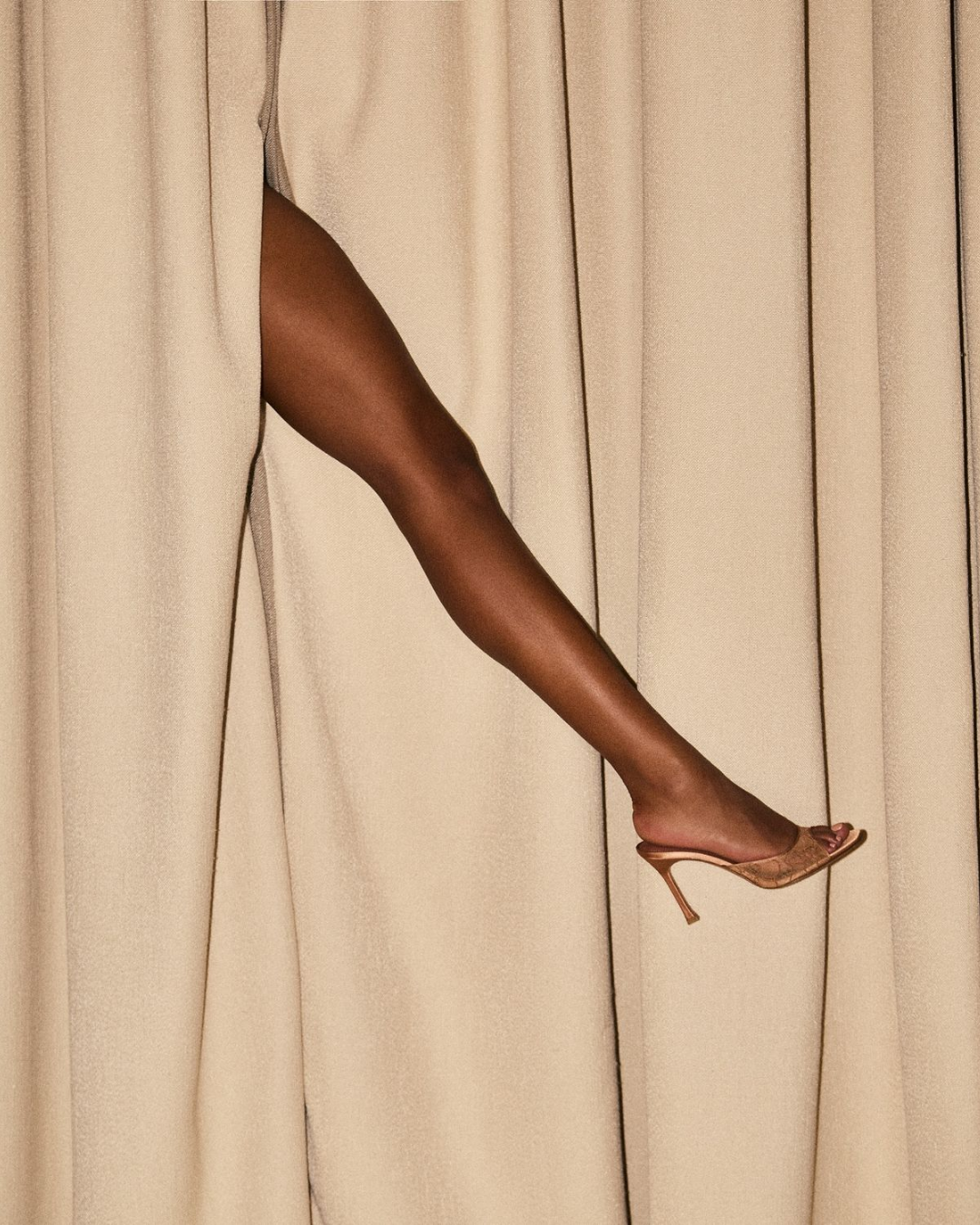 A person's leg in a high heel shoe peeking out from behind beige curtains.
