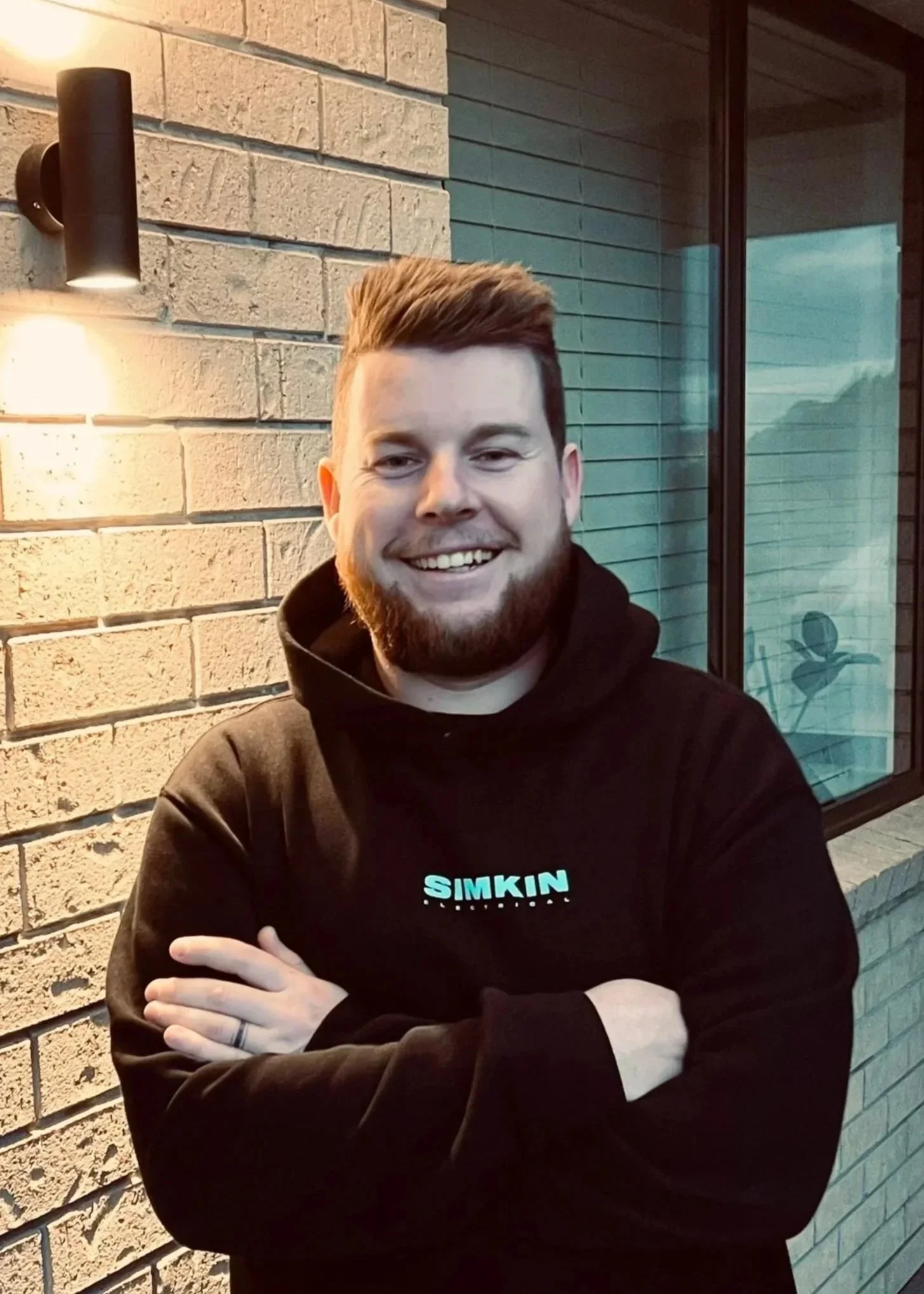 A young man with a beard and styled hair smiling, wearing a black hoodie with the word 'SIMPIN' on it, standing outside near a brick wall and window at dusk.