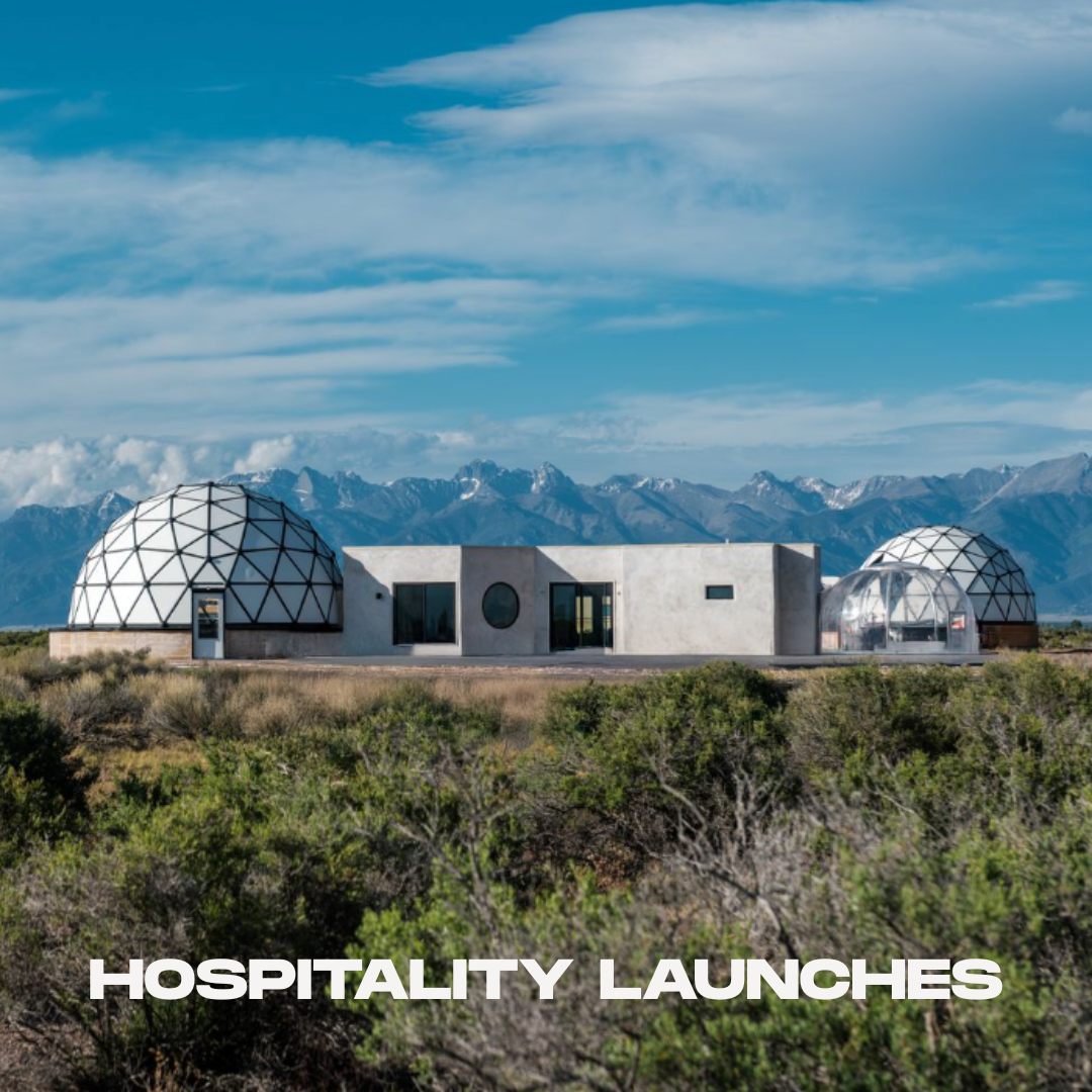 A modern building with geodesic dome structures on a flat landscape with mountains in the background and a partly cloudy sky, with the text 'HOSPITALITY LAUNCHES' at the bottom.