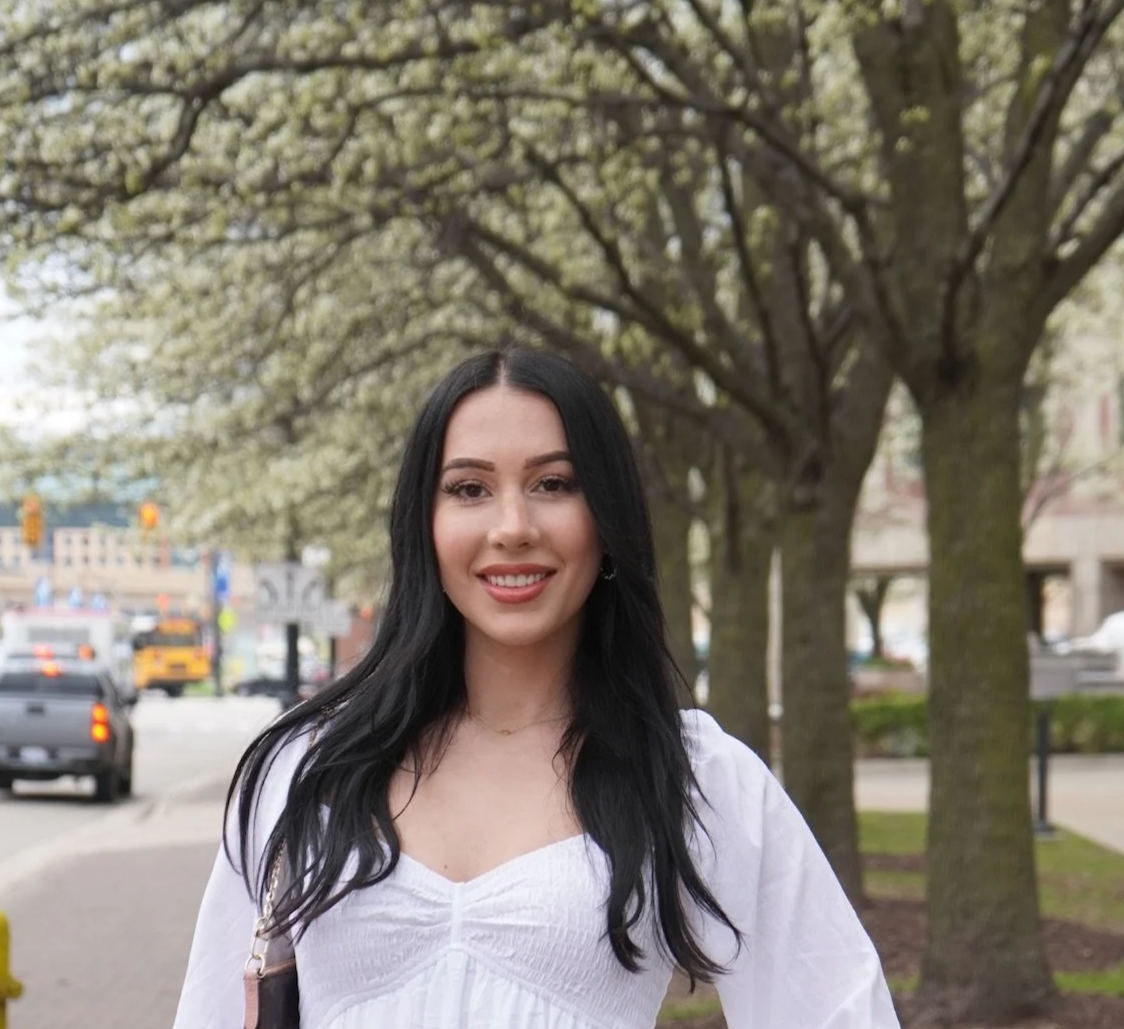 A young woman with long black hair smiling outdoors on a city sidewalk with blooming trees and cars in the background.