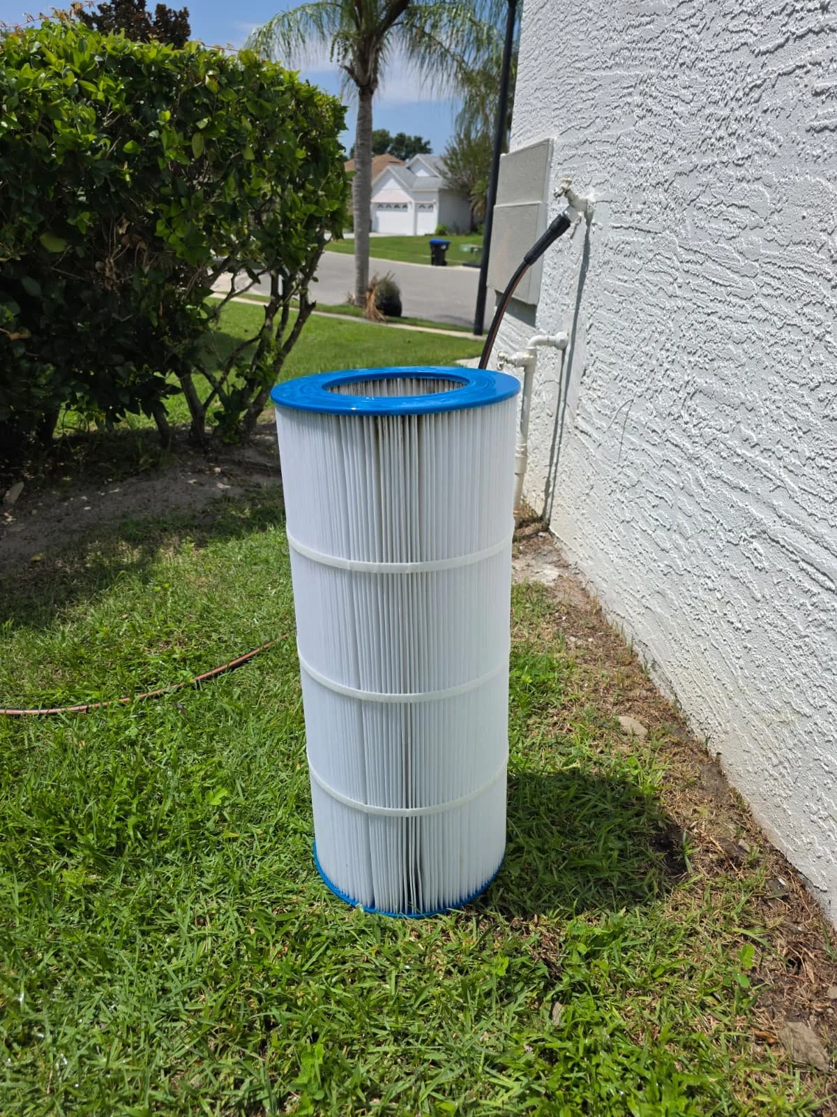 A white pool filter with a blue top standing outdoors on grass next to a white textured wall, with shrubs and a street in the background.
