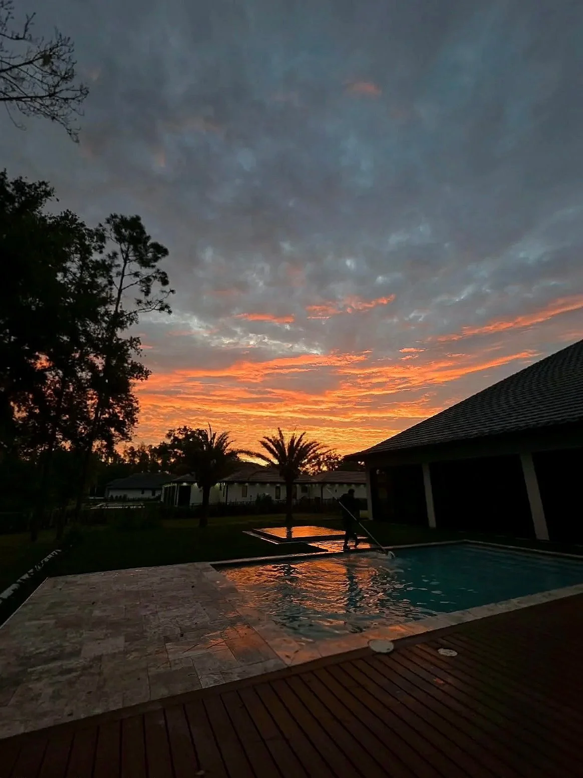 Sunset over a backyard with a swimming pool, trees, and a house. The sky is partly cloudy with orange and pink hues reflecting on the pool water.