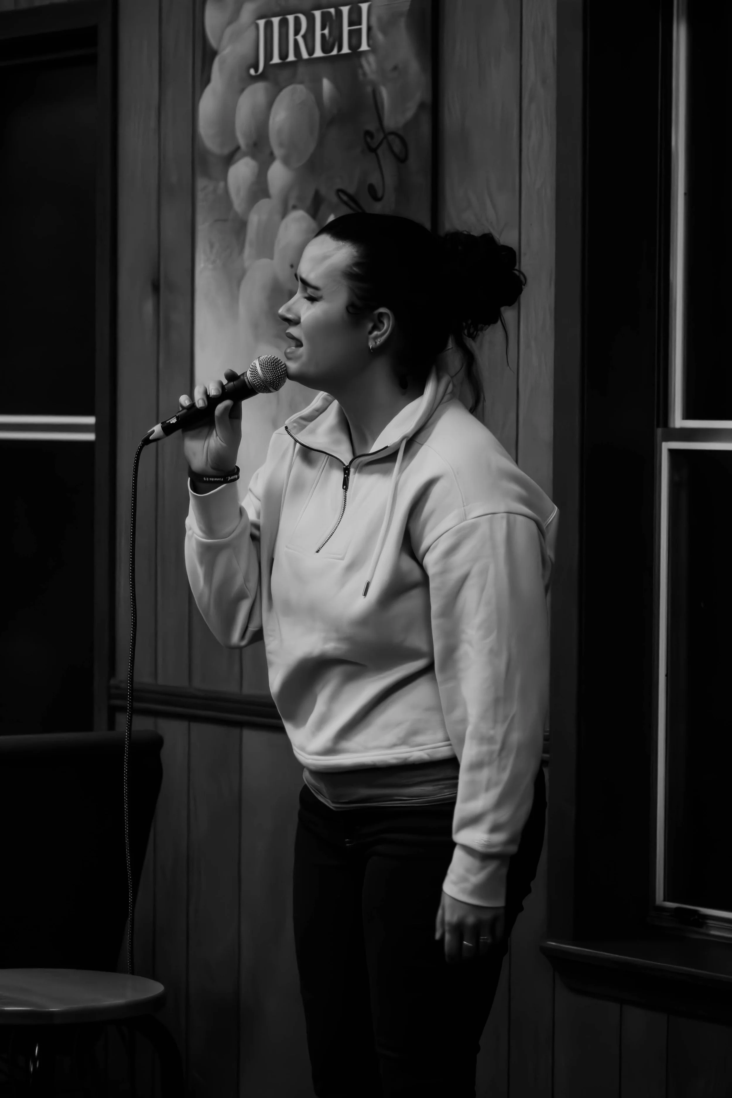 A woman singing into a microphone indoors against wooden paneling wall with a basketball themed sign in the background.