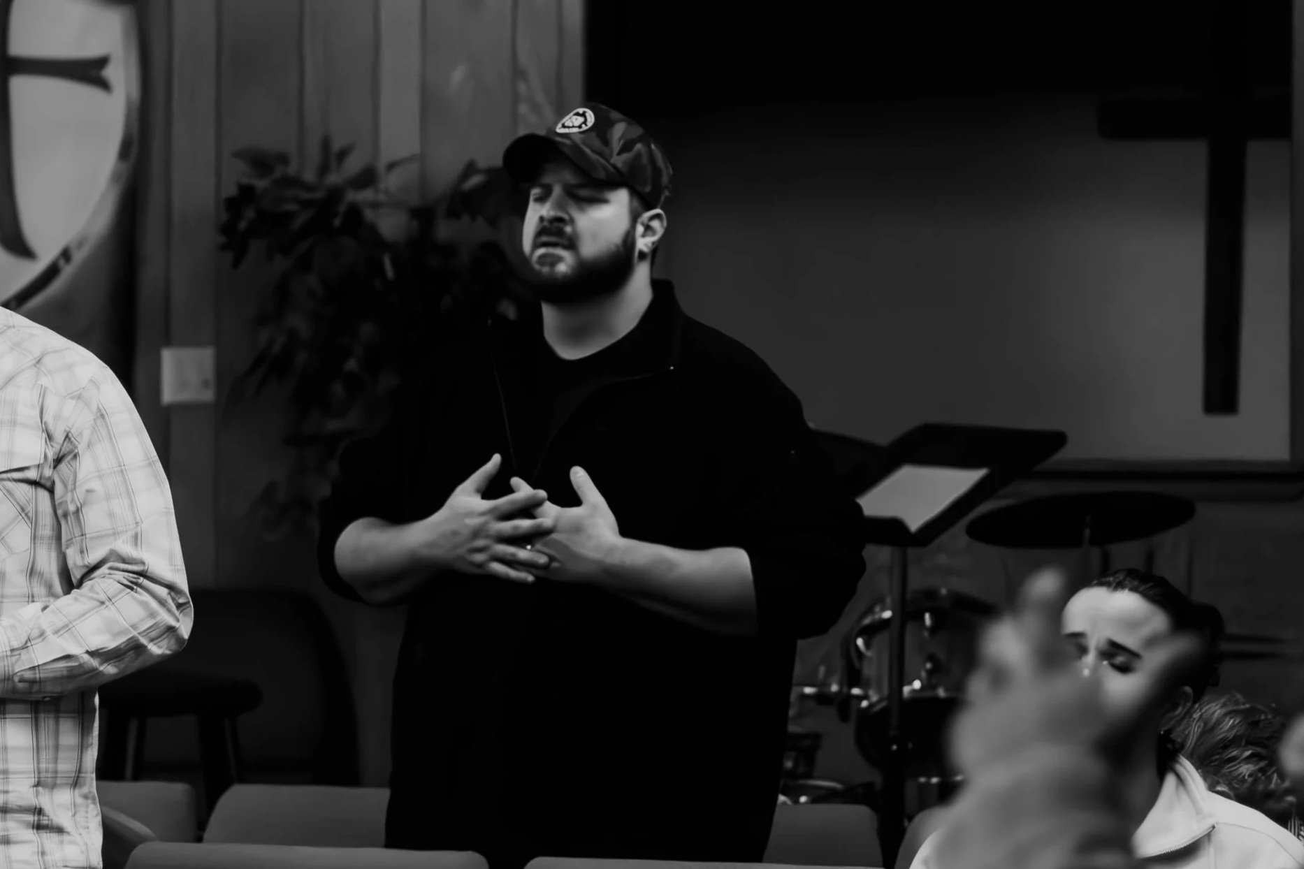 A man with a beard and a cap, standing with hands on chest, appears to be speaking or singing, in a room with wooden walls and musical equipment, including a drum set and sheet music.