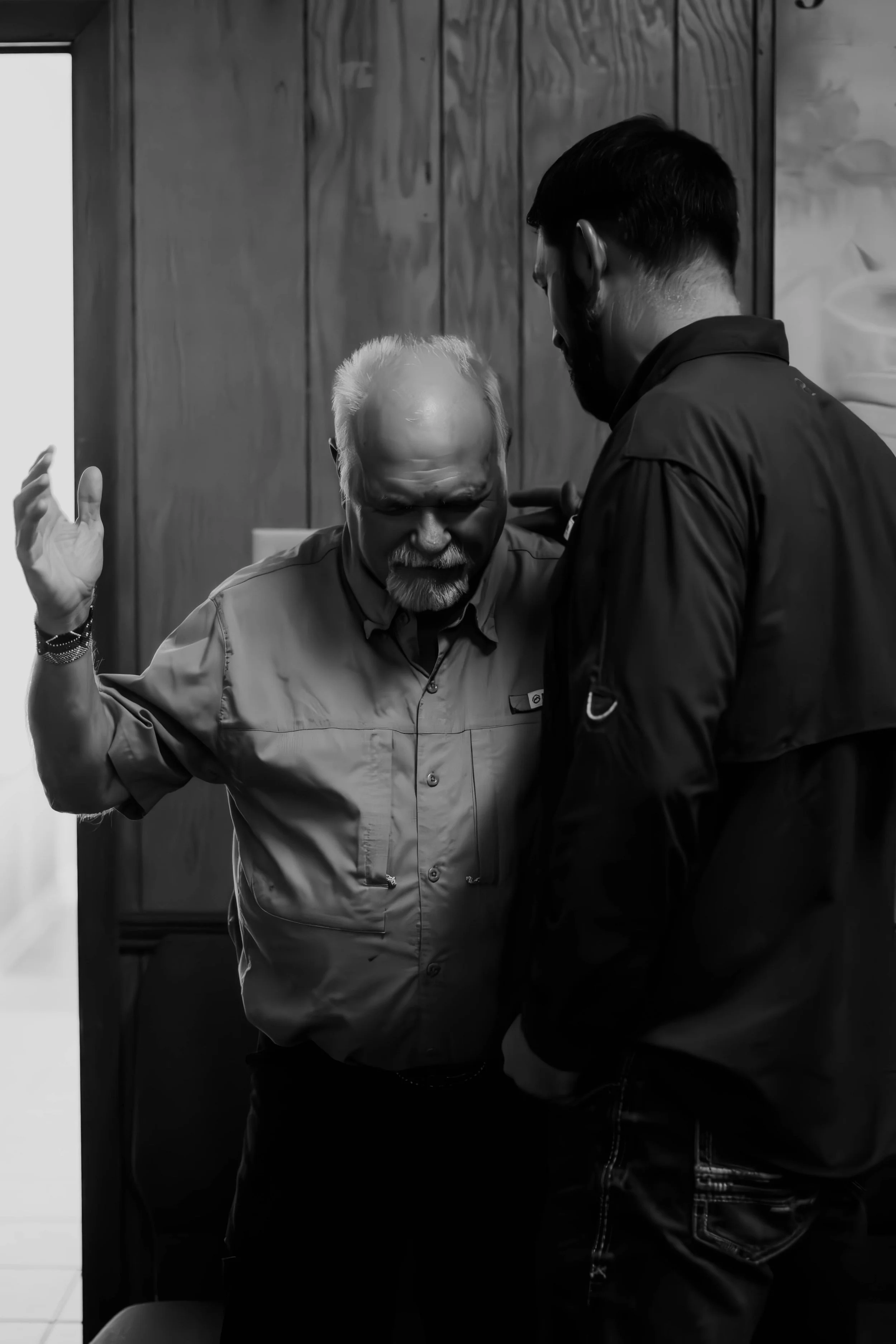 An elderly man with white hair and a beard, raising his hand while talking to a younger man with dark hair, in a room with wooden paneling.