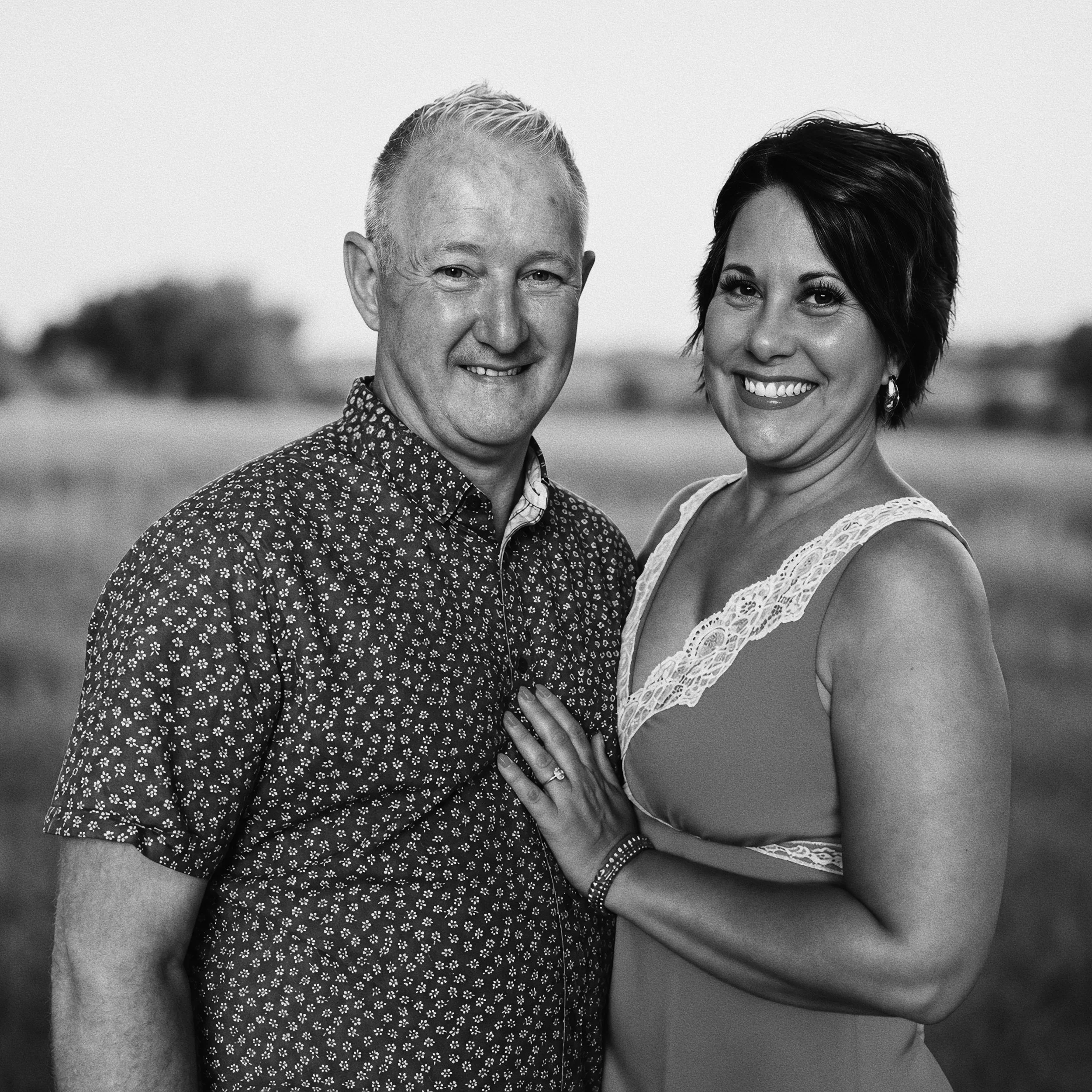 Close-up of a smiling couple, a man with short, light hair and a woman with short dark hair, standing outdoors with a blurred natural background.