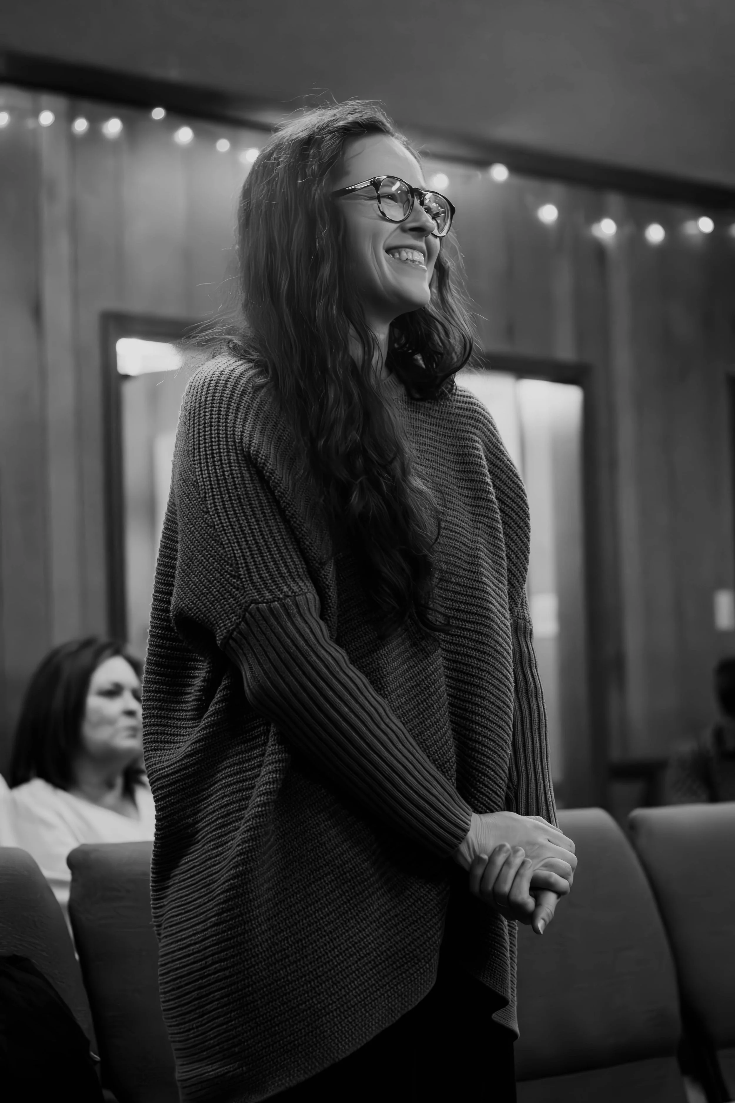 A woman with glasses, long curly hair, and a sweater smiling and standing with her hands clasped in a room with wooden walls and string lights, while other people sit nearby.