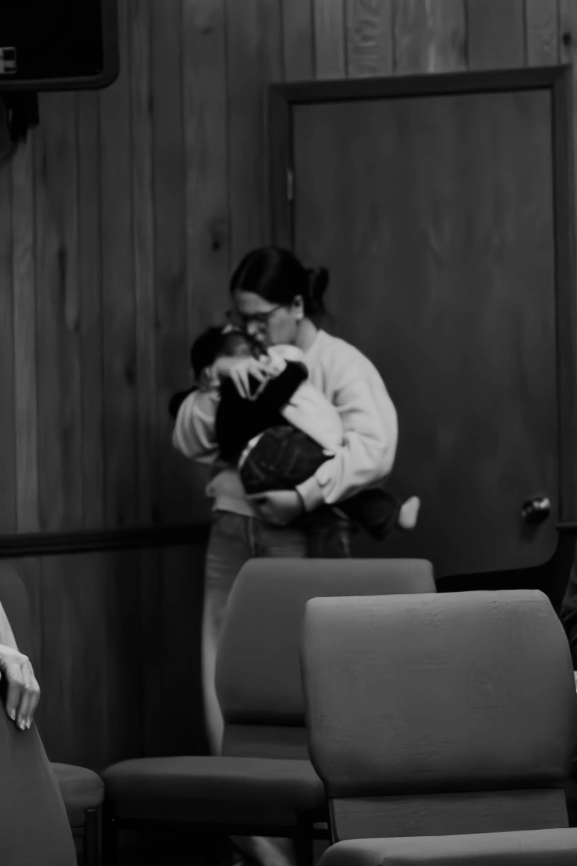 A woman in glasses holding a small dog in her arms while sitting at a table in a room with wooden panel walls, with chairs in the foreground and a mirror on the wall.