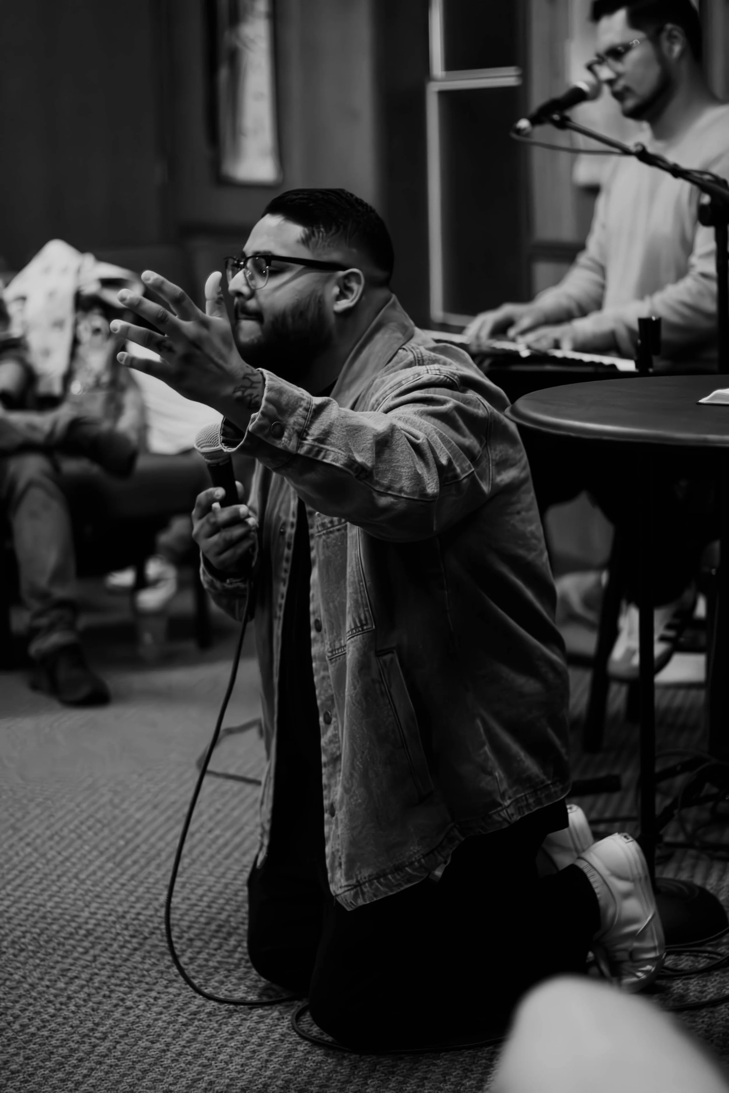 A person kneeling on the floor with eyes closed, holding a microphone in one hand and raising the other hand with fingers spread. They are wearing glasses and a denim jacket. In the background, another person is playing a keyboard and singing into a microphone. The setting appears to be an indoor room with wood-paneled walls.