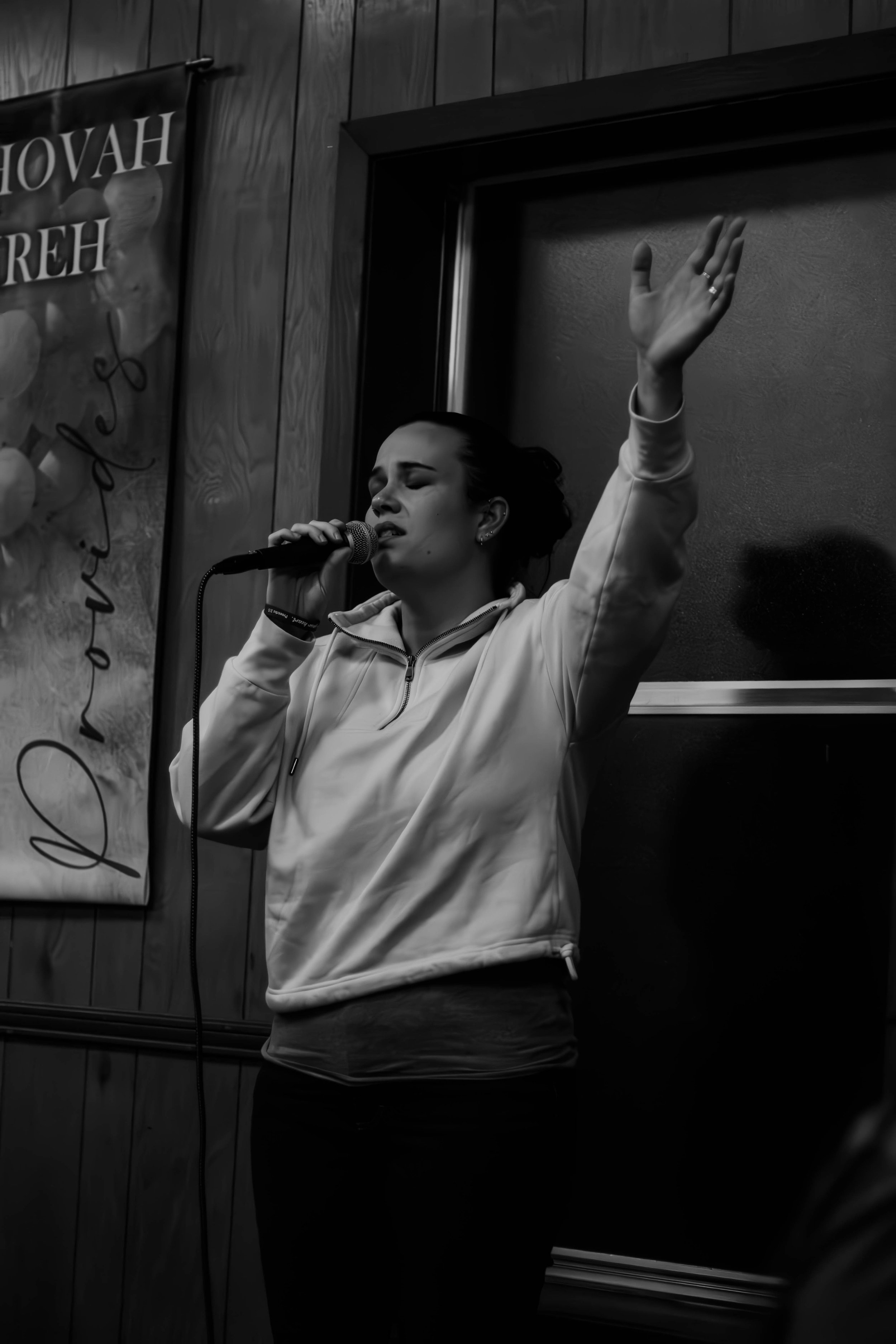 A young woman singing passionately into a microphone, with her eyes closed and one hand raised, standing indoors in front of a wooden wall with a partial banner on the side.