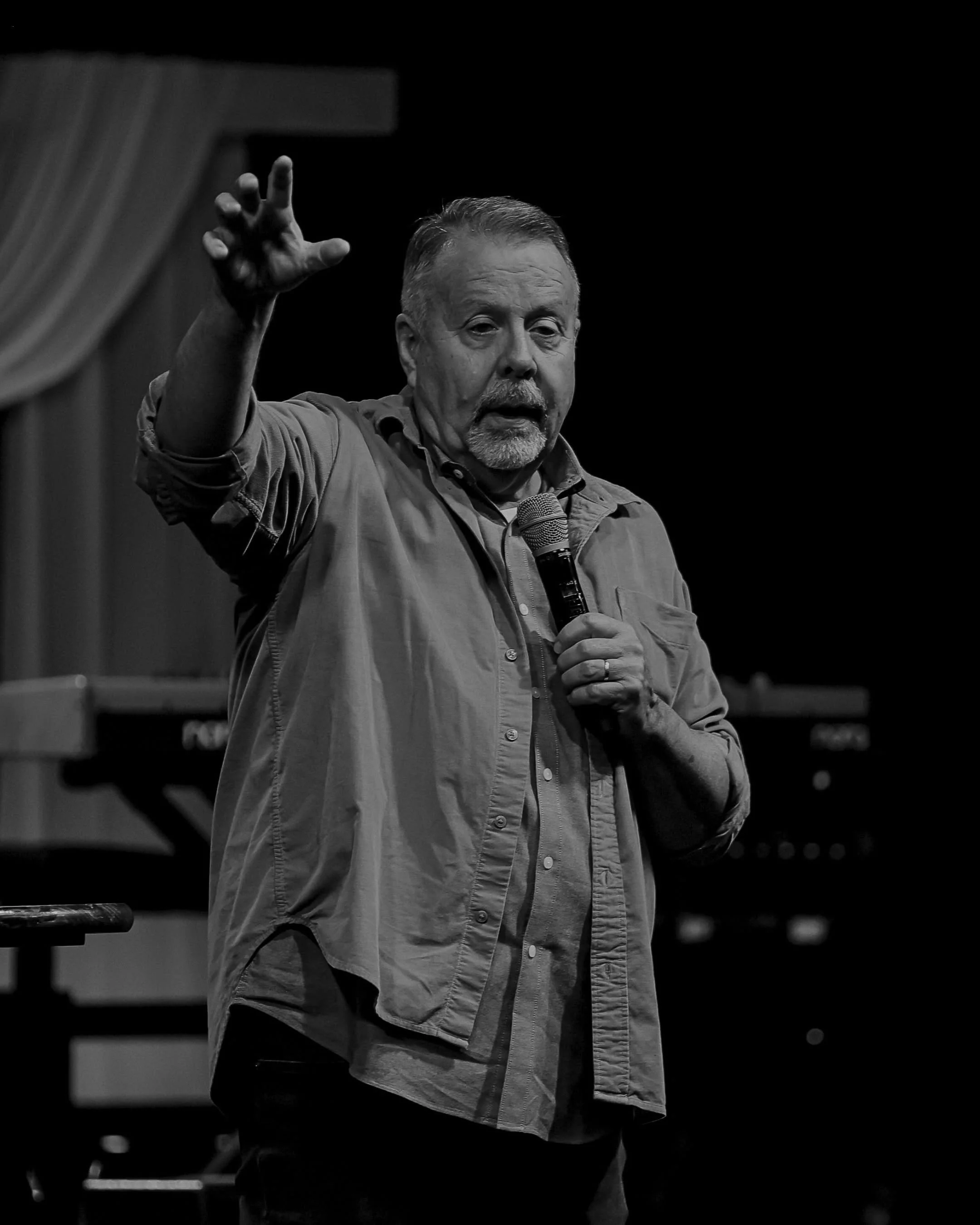 A middle-aged man with short hair and a beard is speaking into a microphone while gesturing with his right hand during a presentation or speech in a dimly lit room.