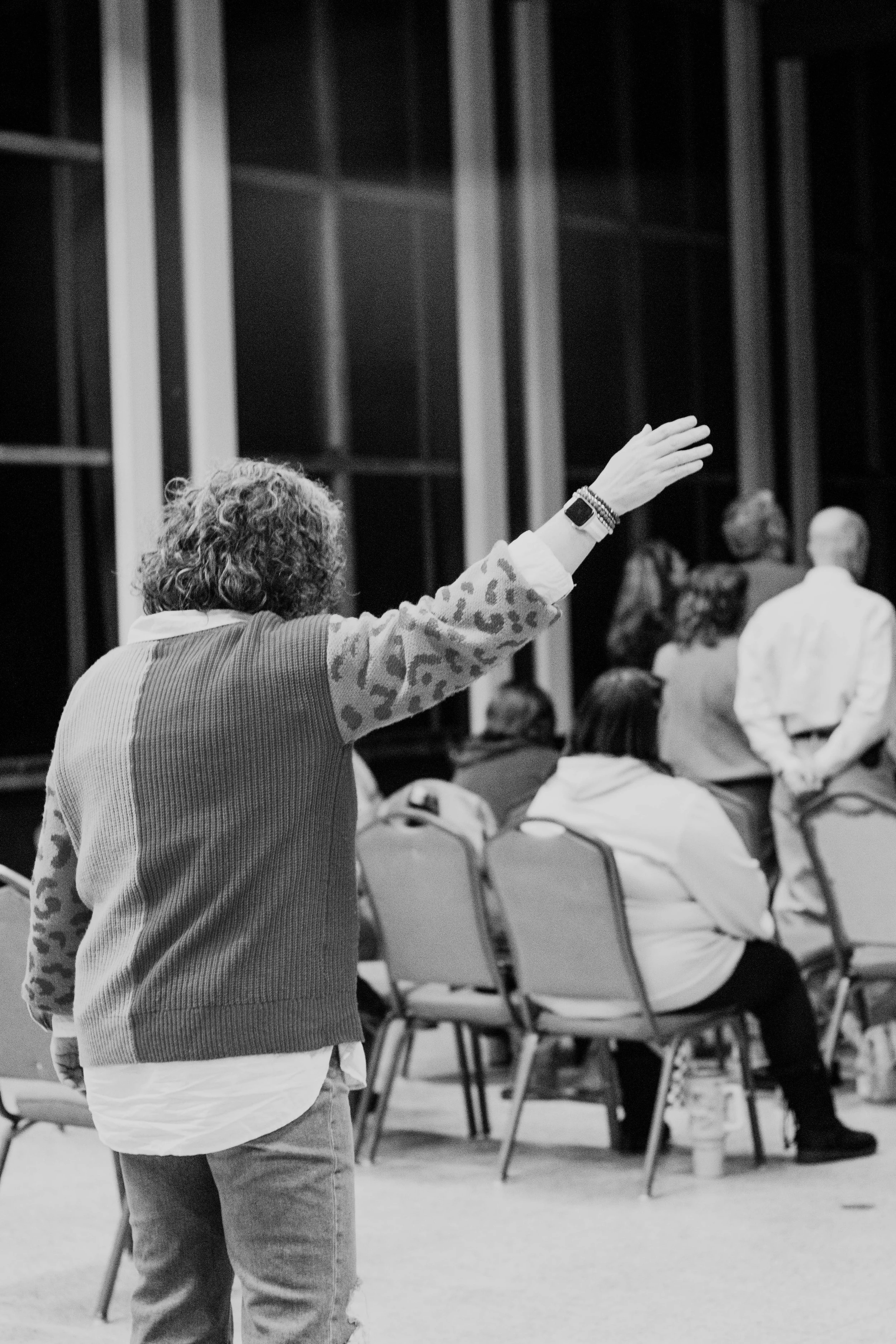 Woman standing in a room with chairs and people, raising her hand, with a large window in the background.