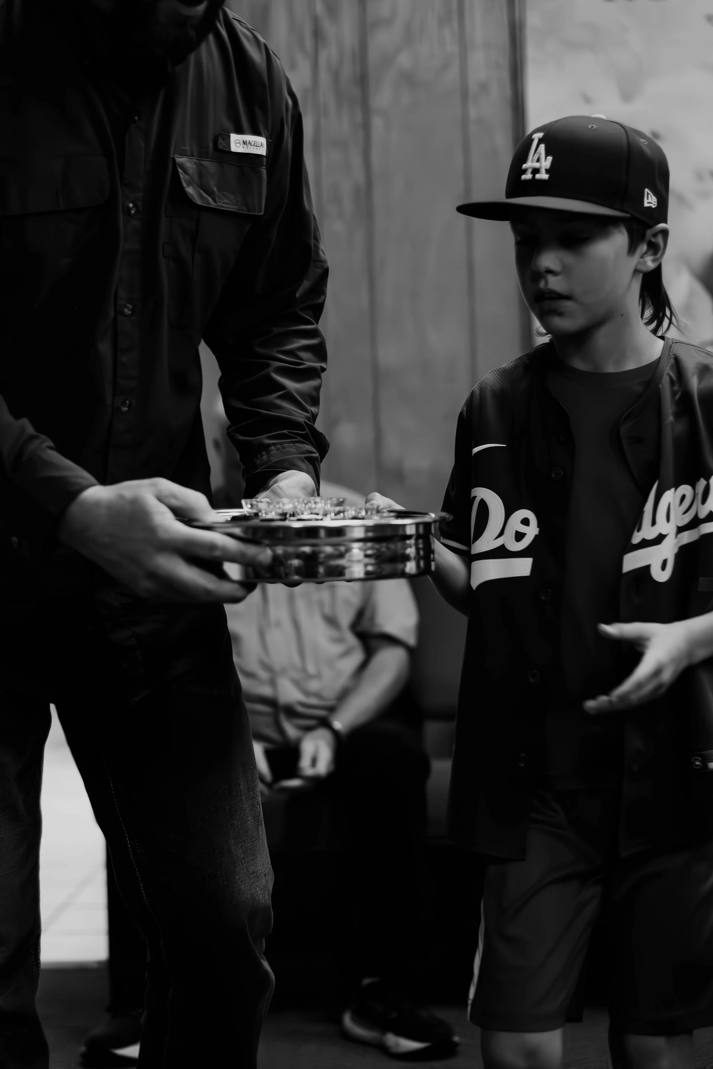 A young boy wearing a Los Angeles Dodgers baseball cap and jersey, standing with a person in a dark uniform handing him an object, in black and white.