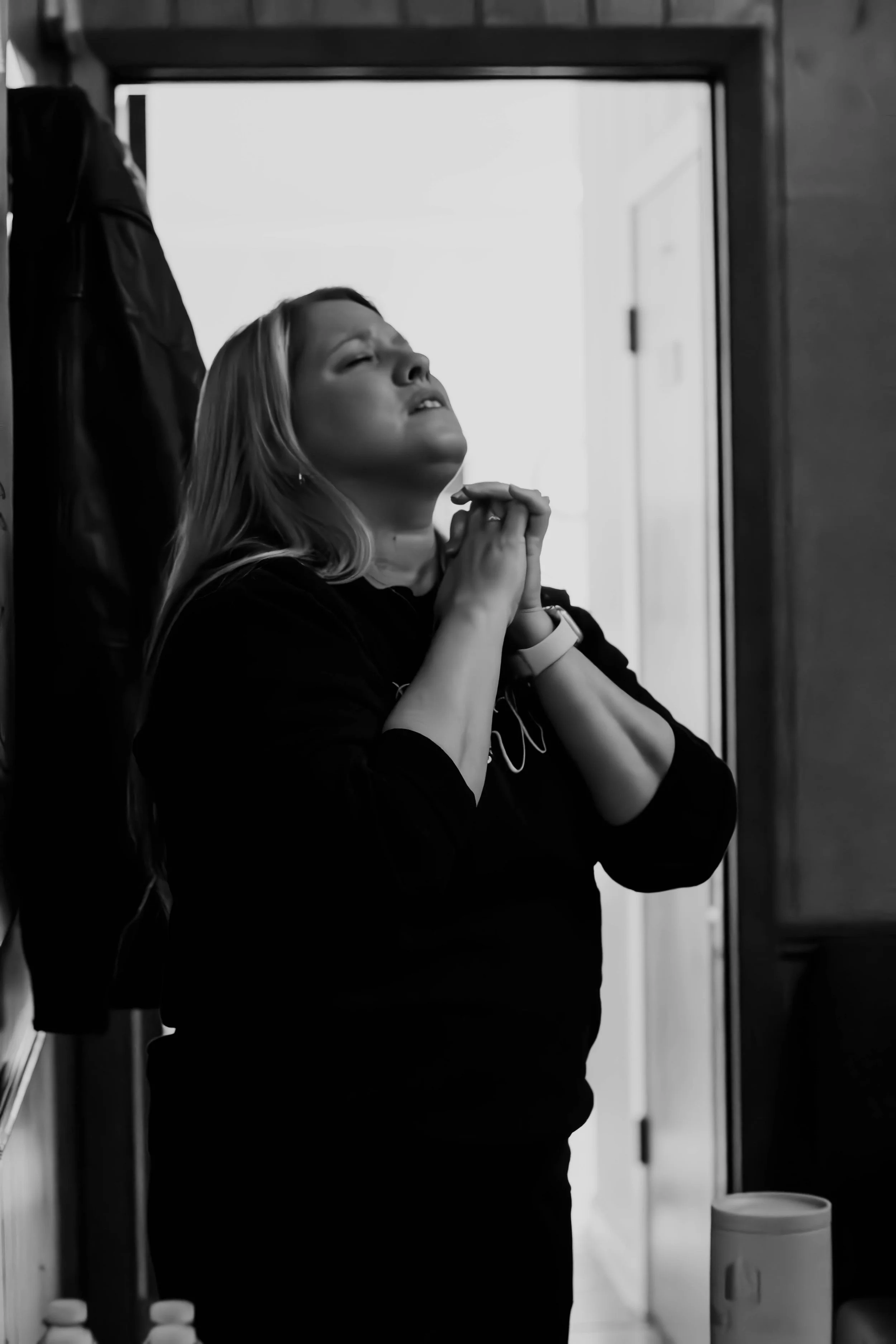 A woman with long hair and a watch on her wrist is praying with her eyes closed and hands clasped in front of a doorframe in a kitchen.