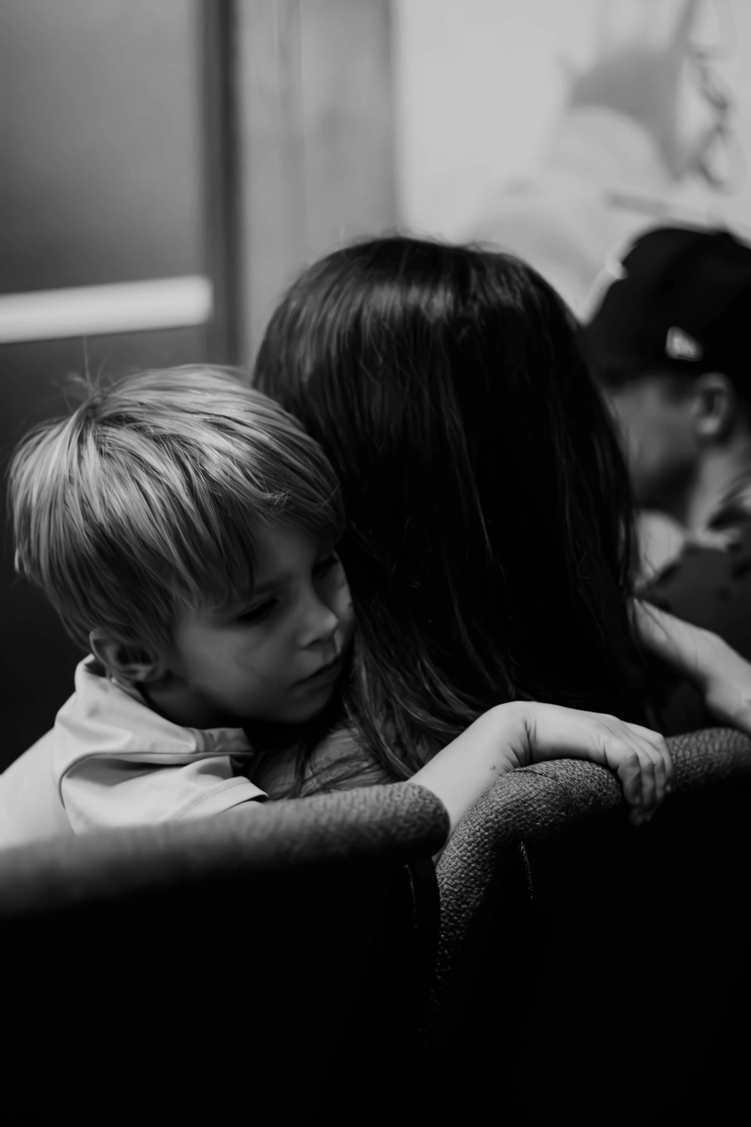 A grayscale photo of two children hugging each other, with one child having blonde hair and the other having dark hair. They are sitting in a space that appears to be a classroom or similar environment.