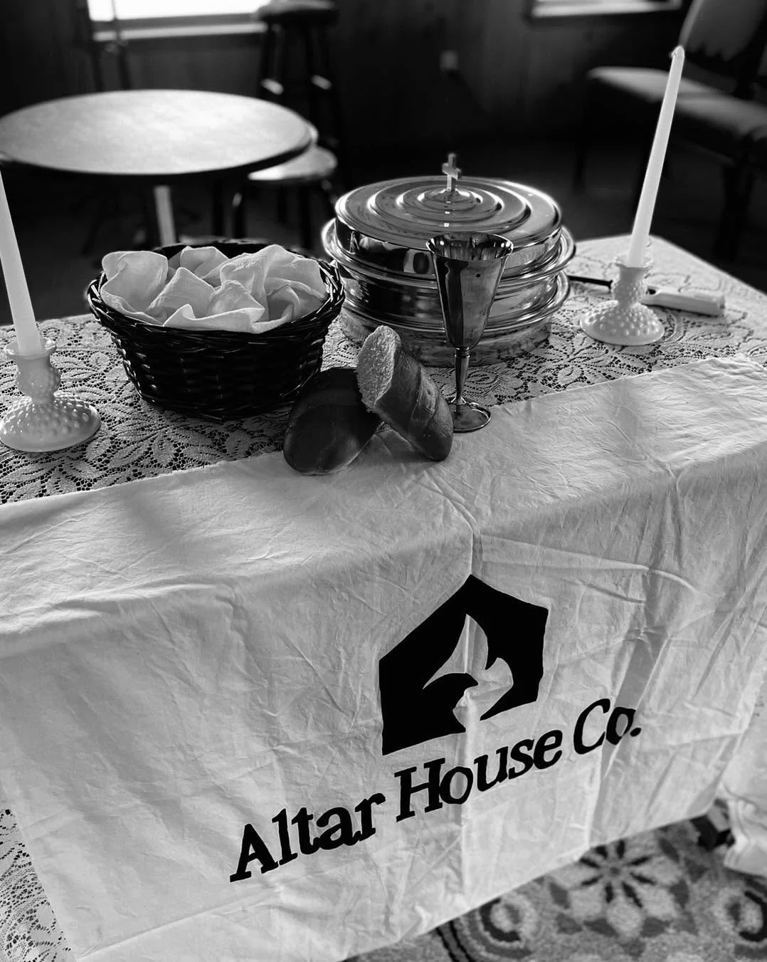 A table set for a religious or ceremonial event with a cloth bearing the Altar House Co. logo. The table holds two lit candles, a basket of tissue paper, a glass bowl with a lid, a cup, and some bread or rolls.