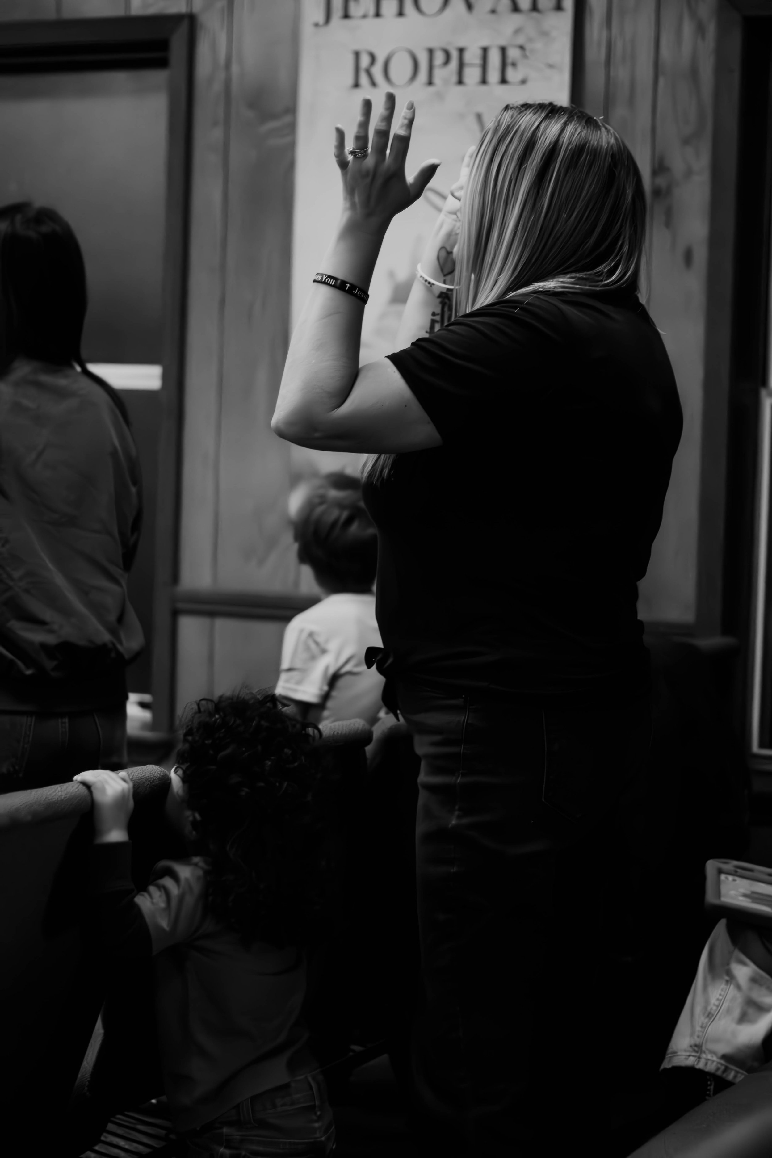 A woman with long hair and a face mask is standing with her hand raised, facing a poster in what appears to be a classroom or educational setting. There are children sitting in front of her, some leaning on a railing. The environment looks like a school or an auditorium.