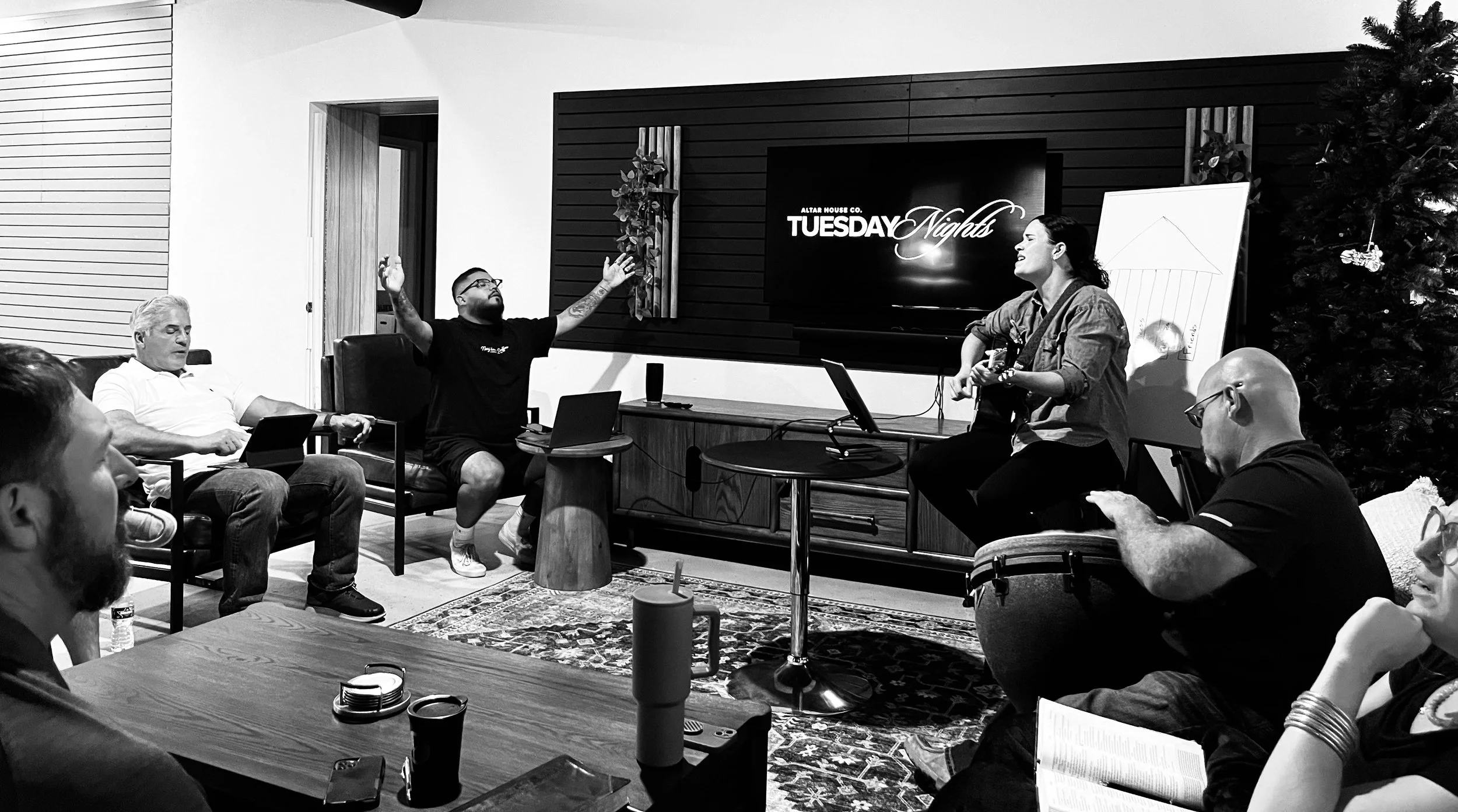 A group of people participating in a meeting or workshop in a cozy, modern room. There is a woman playing guitar, a man with his arms raised, a large screen displaying 'Tuesday Nights,' and a decorated Christmas tree in the background.