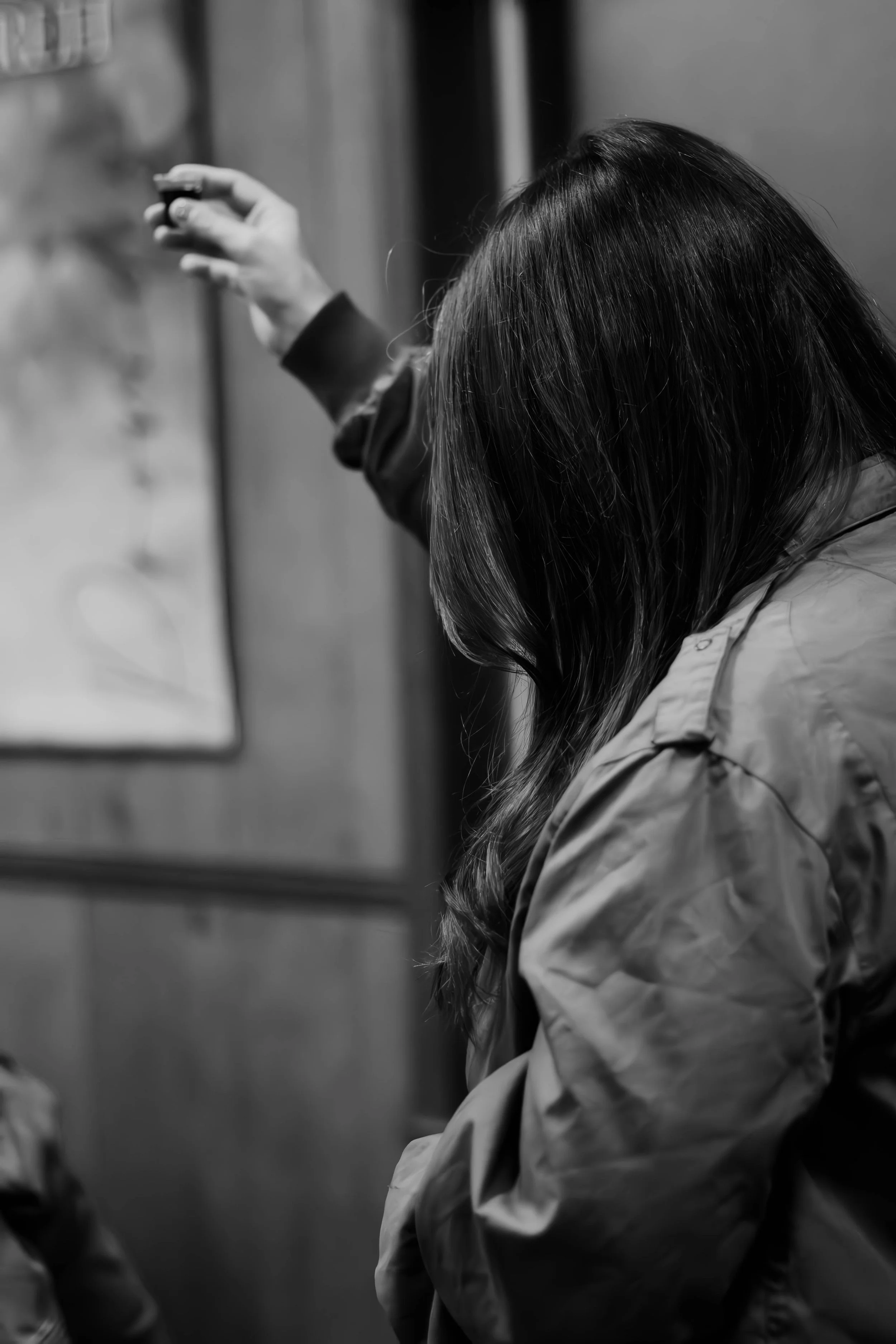 A woman with long hair, wearing a jacket, tilts her head downward and is seen from the side. She is in an indoor setting, possibly an elevator, with blurred background details.