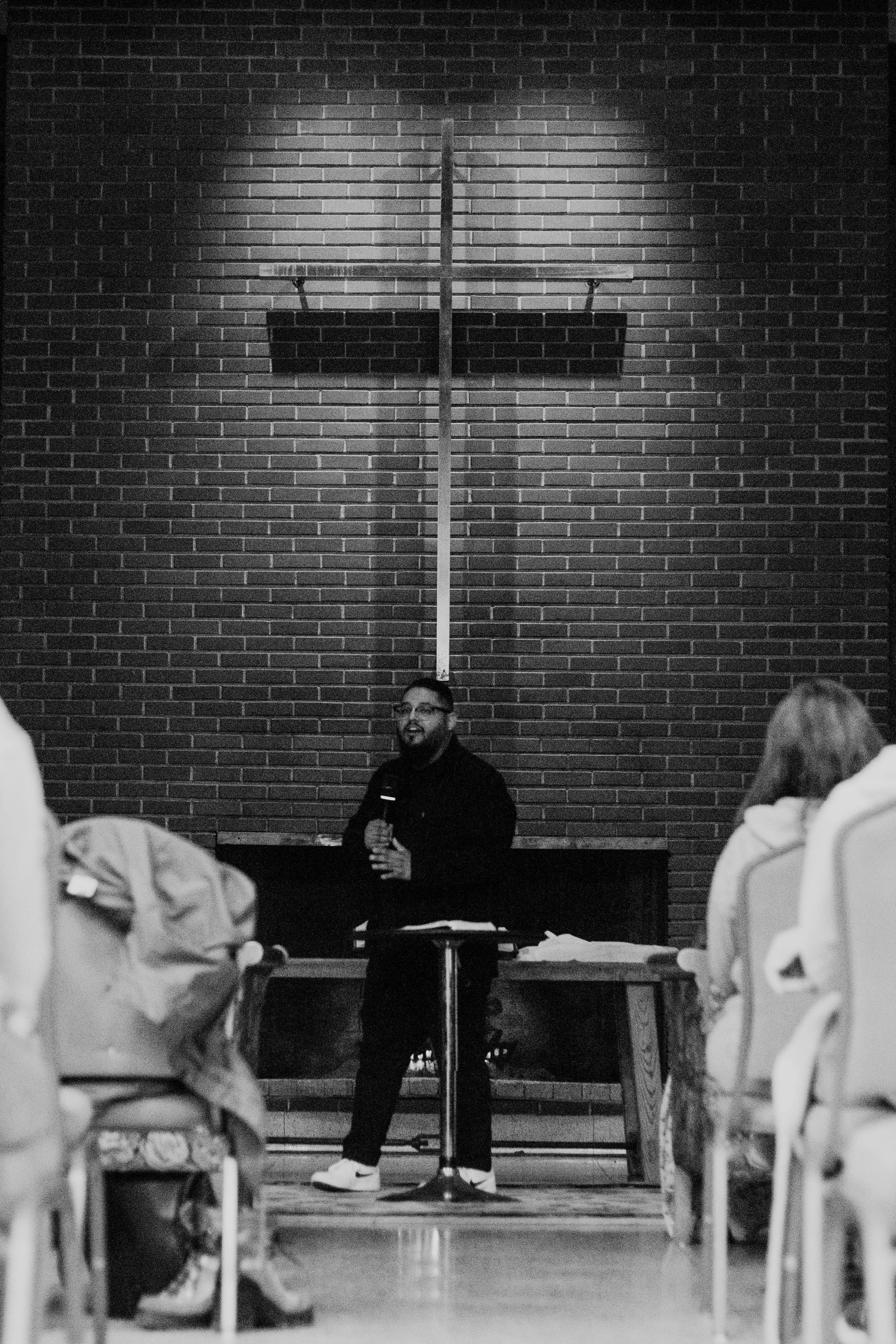A man giving a speech or presentation in front of a brick wall with a cross. Several people are seated at tables facing him, listening.