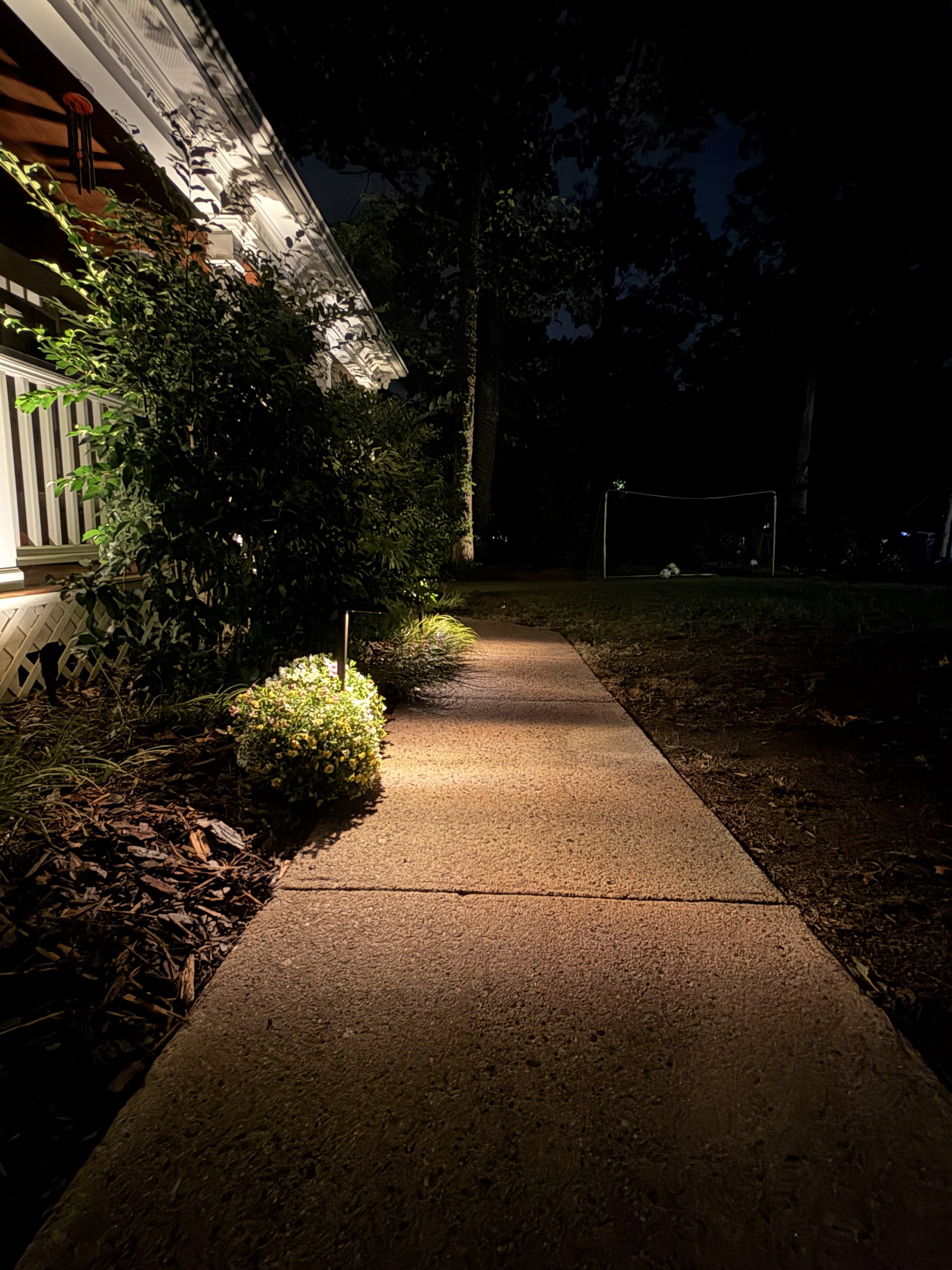 Nighttime scene of a sidewalk next to a house with a porch. The porch is illuminated, and a garden with bushes and flowers is visible. In the background, a soccer goal and some soccer balls are seen in the yard.