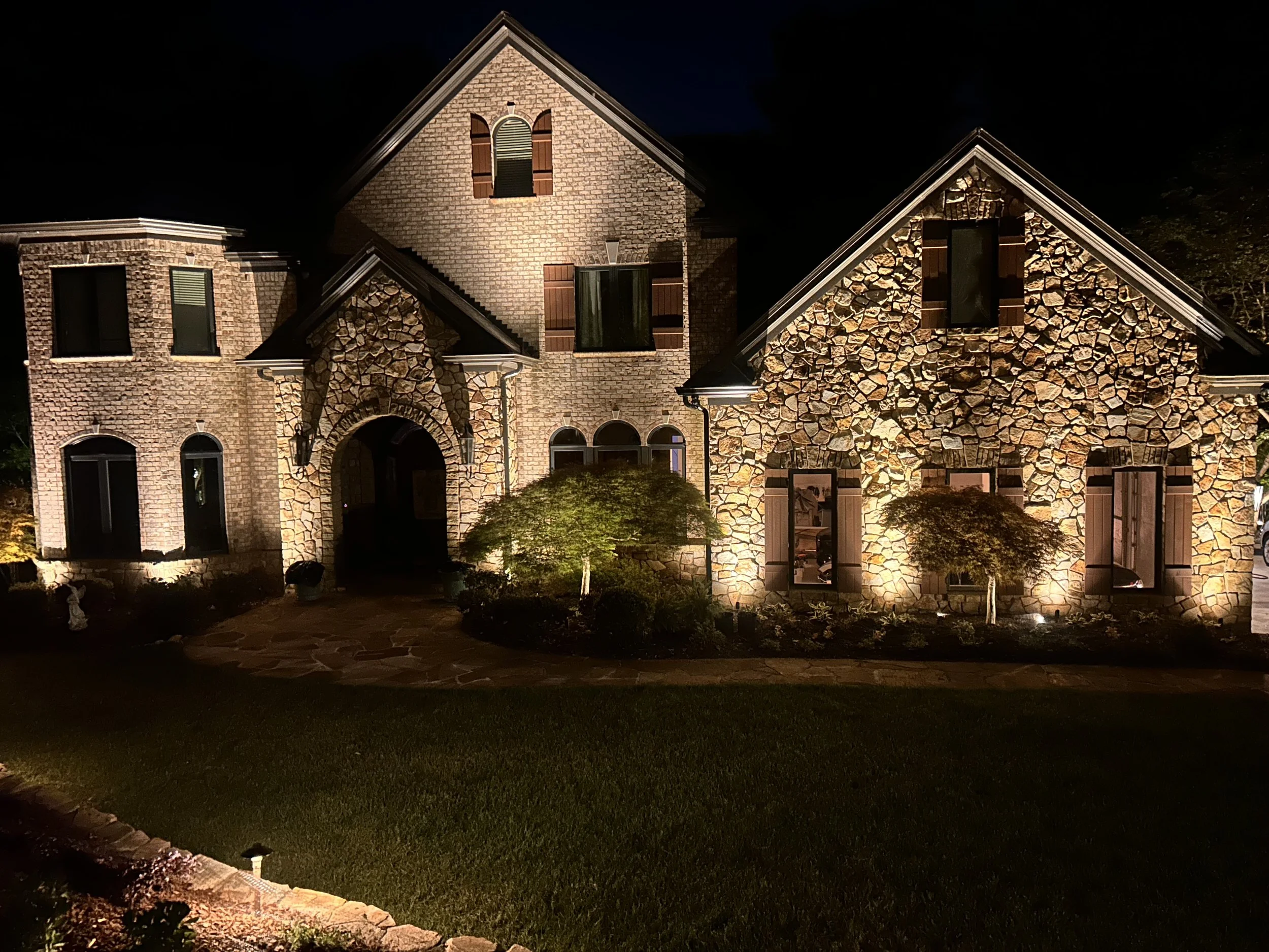 A large house at night with brick and stone exterior, lit outdoor lighting, and a well-maintained front yard.