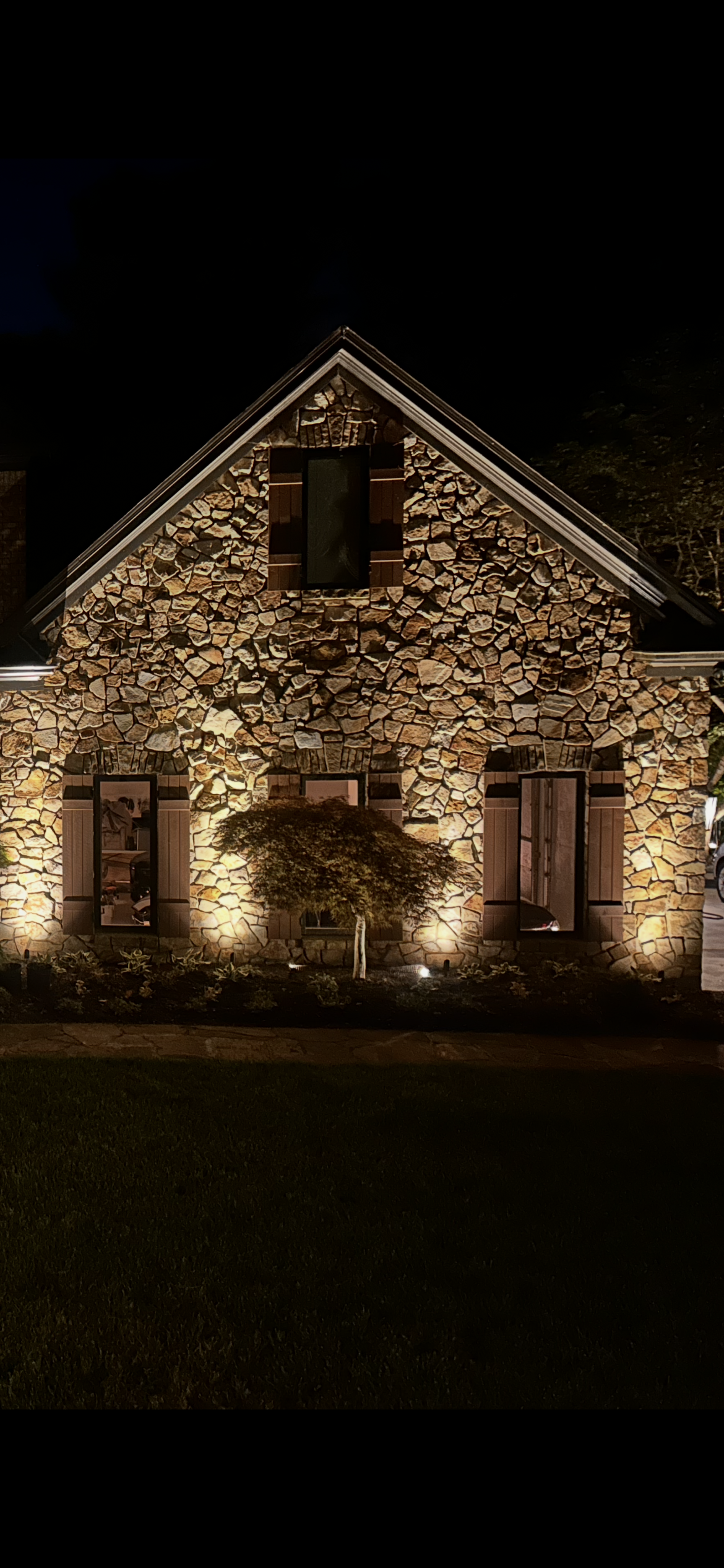 A stone house at night with warm exterior lighting, three windows with wooden shutters, and a small tree in front.