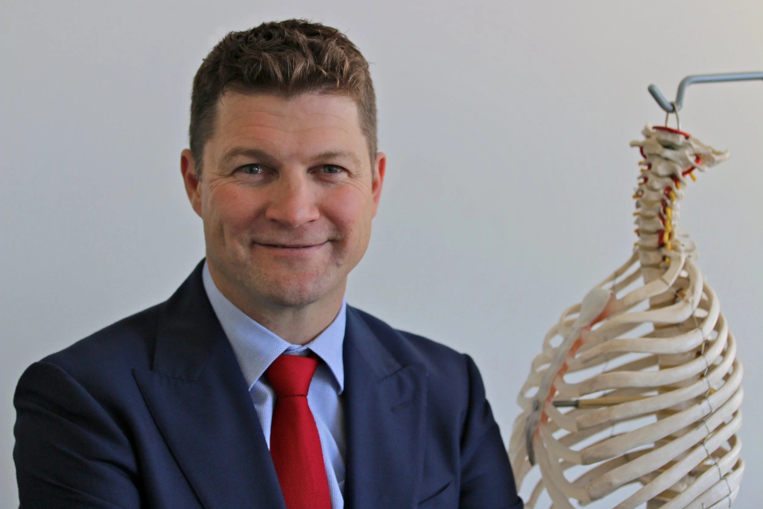 A man in a business suit with a red tie smiling, next to a human skeleton model.
