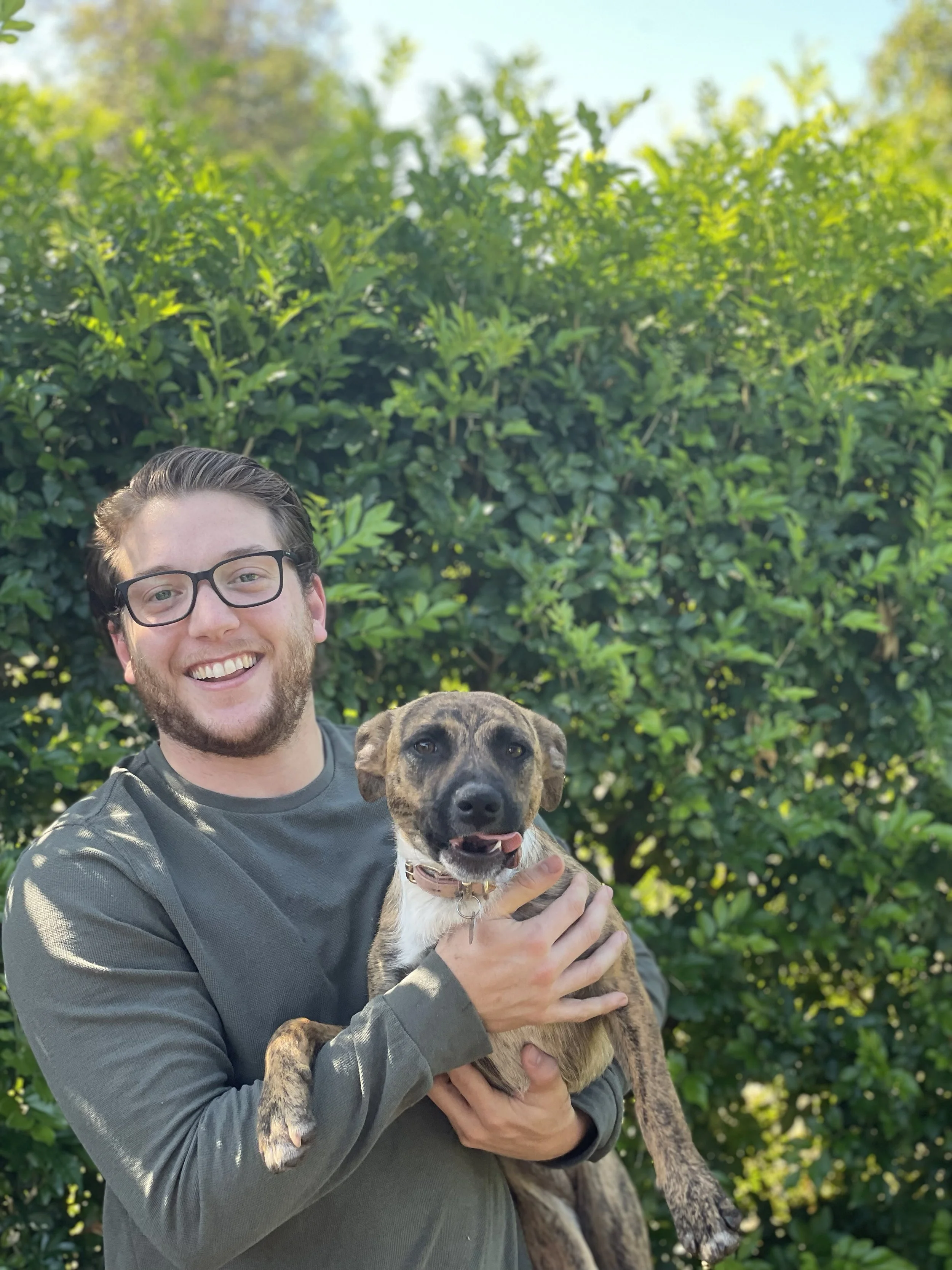 A man with glasses smiling while holding a brown and black dog with a playful expression outdoor with greenery in the background.