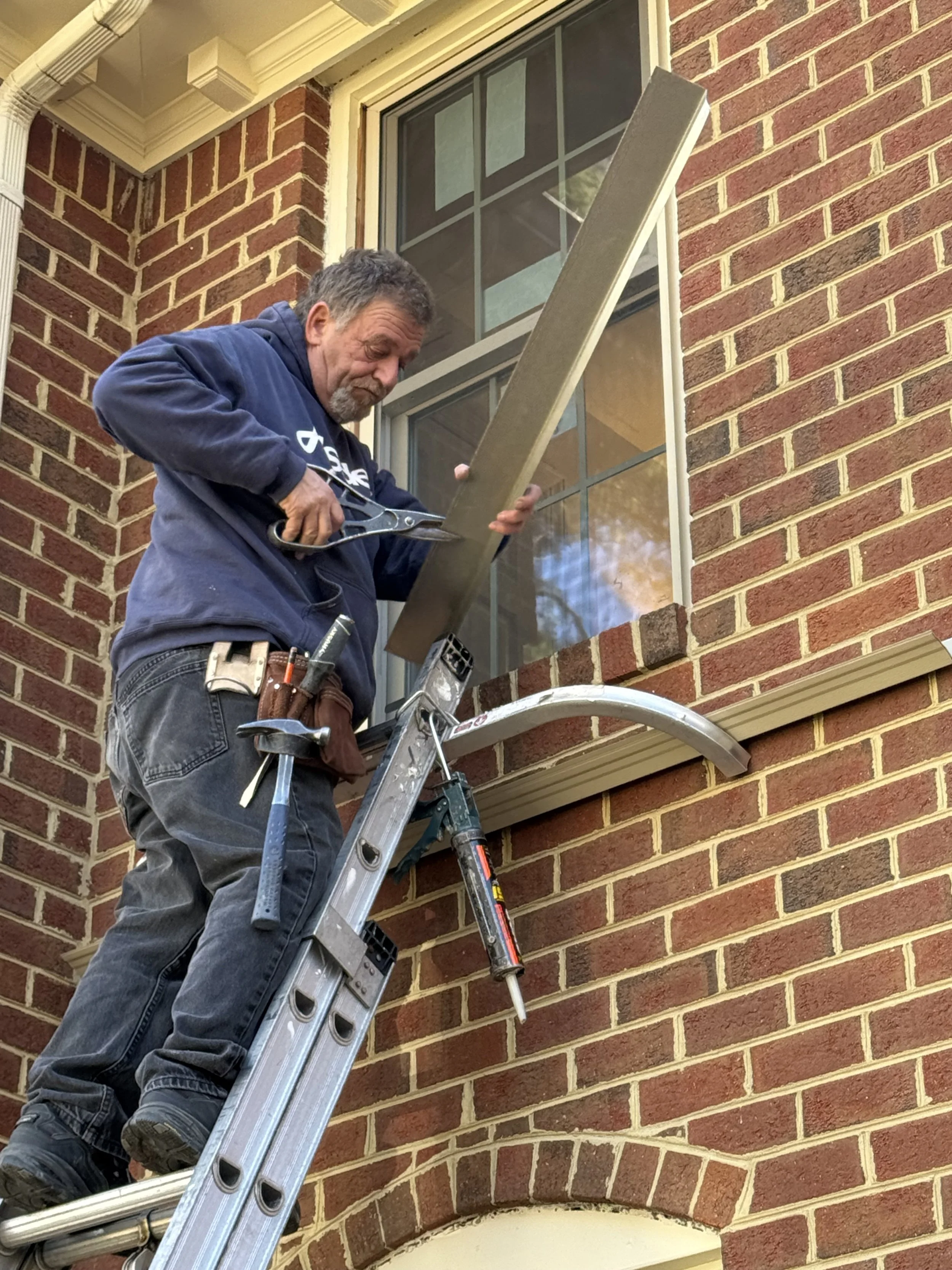 A man climbing a ladder outside a brick building, holding a long wooden plank, with tools attached to his belt, preparing to work on the building's exterior or window.
