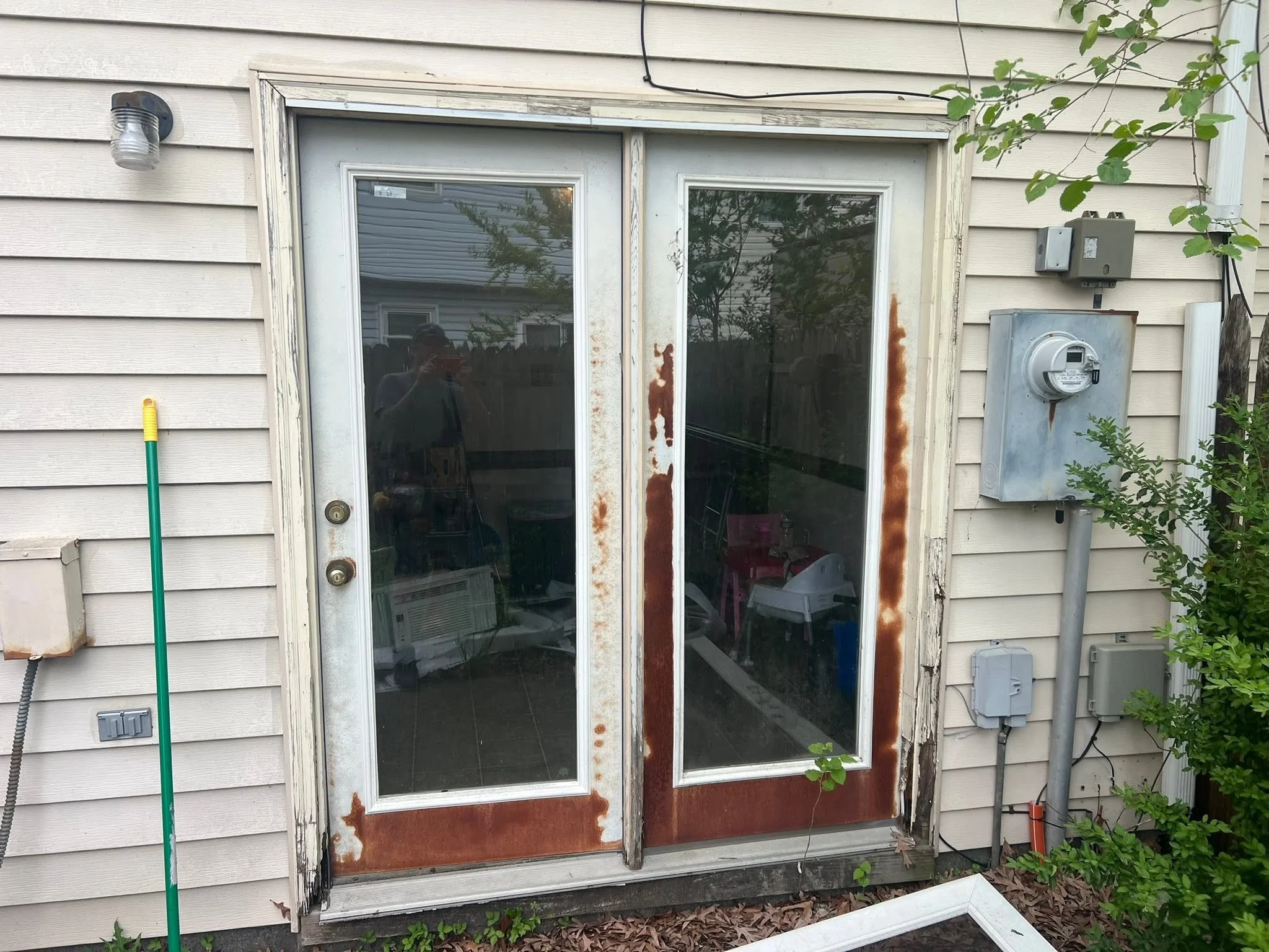 A sliding glass door with rust and damage along the edges, set in a house with beige vinyl siding, surrounded by some green plants and a yellow-handled gardening tool leaning against the wall.