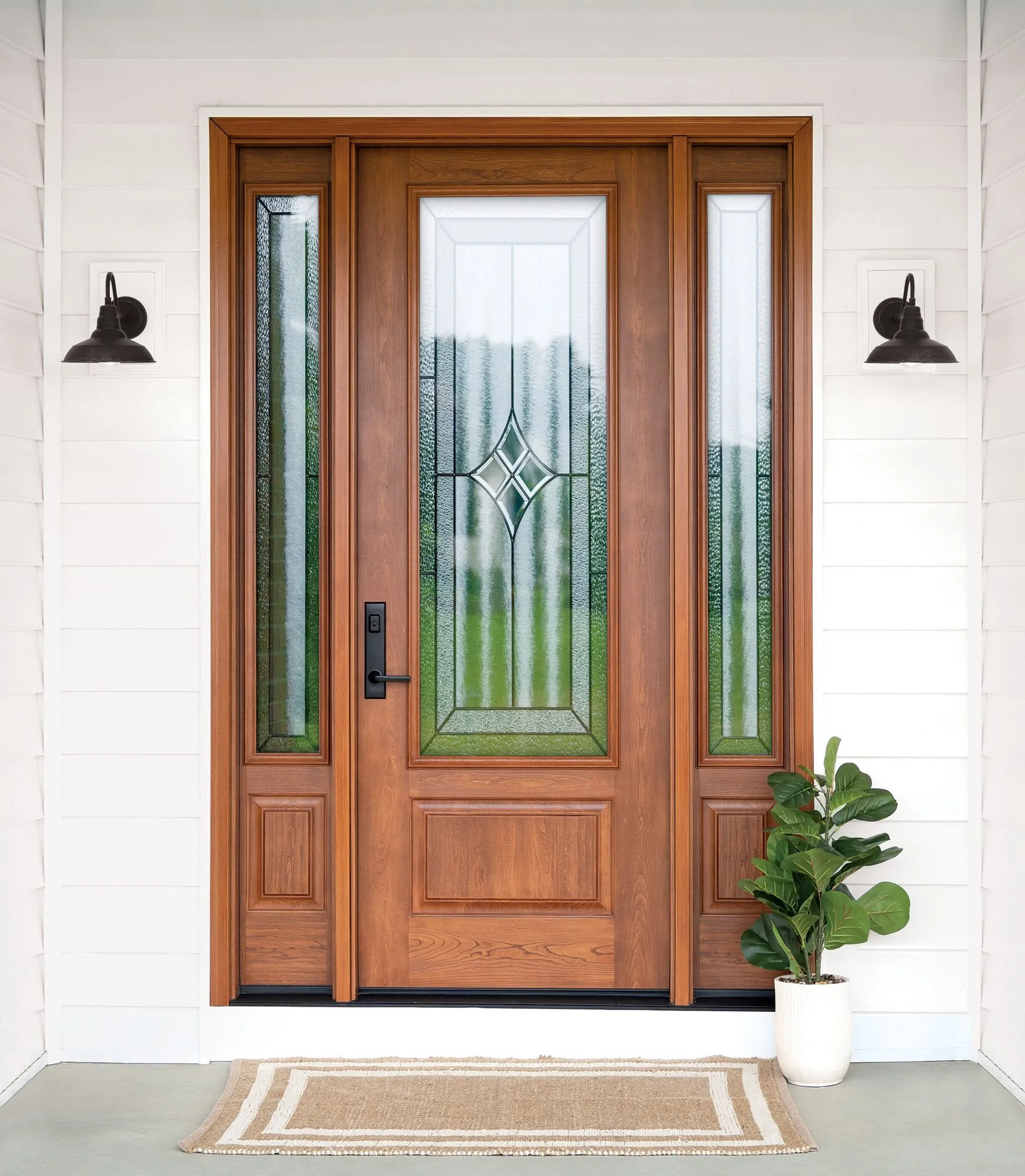 A front door with a wooden frame and decorative glass panels, flanked by wall-mounted black outdoor lights, with a potted plant and a beige rug outside.