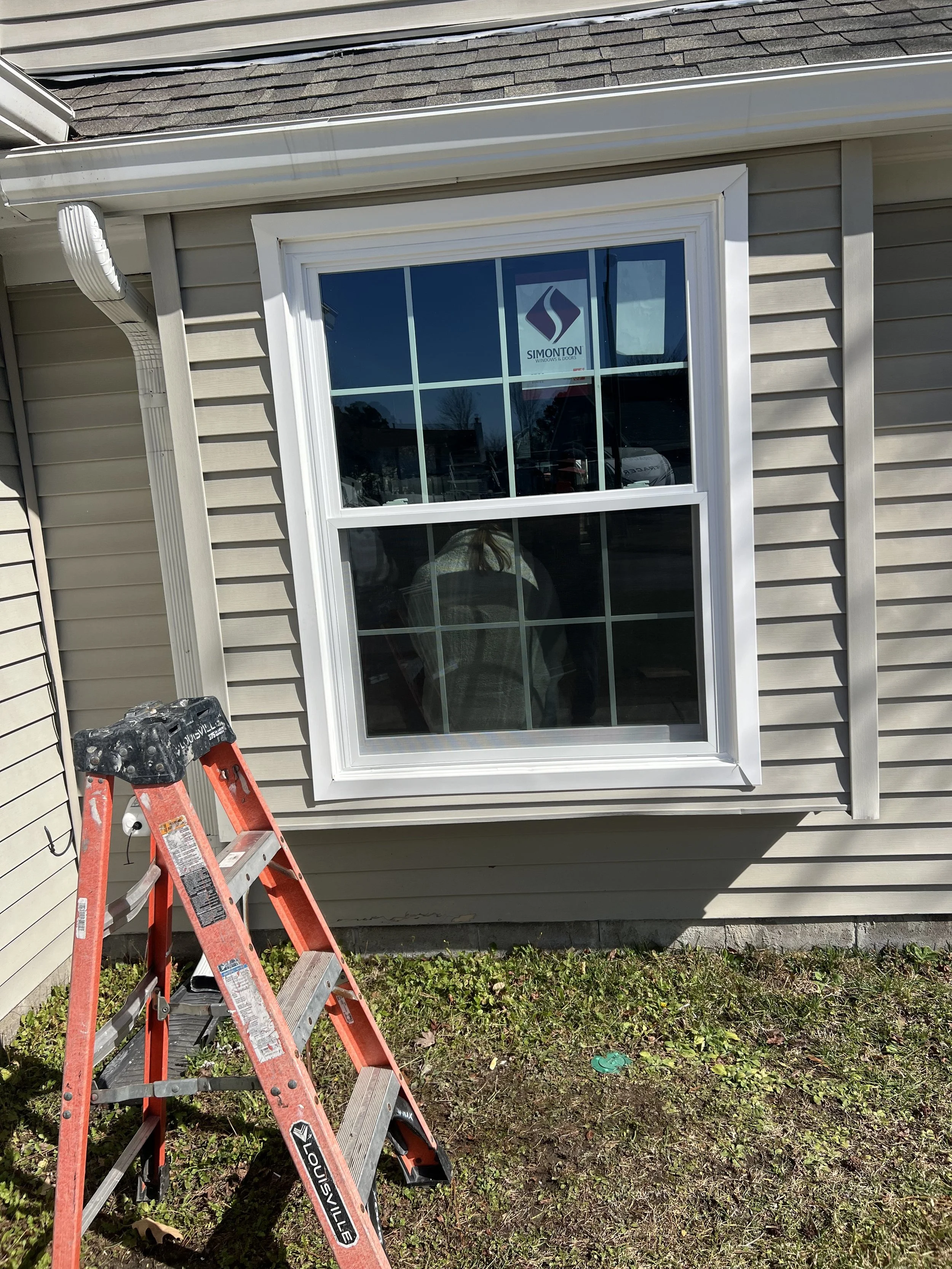 A closed window on the beige siding of a house with white framing. There is a gray gutters and downspout system nearby. A red step ladder is positioned in front of the window, on a grassy area with patches of dirt and small plants.