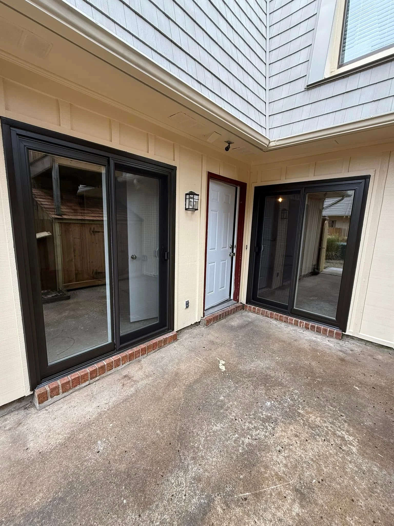 Backyard patio area with two sliding glass door entrances, a white door with a red frame, and an exterior wall with beige siding and gray shingles above. The ground is concrete with loose dirt and patches of gravel.