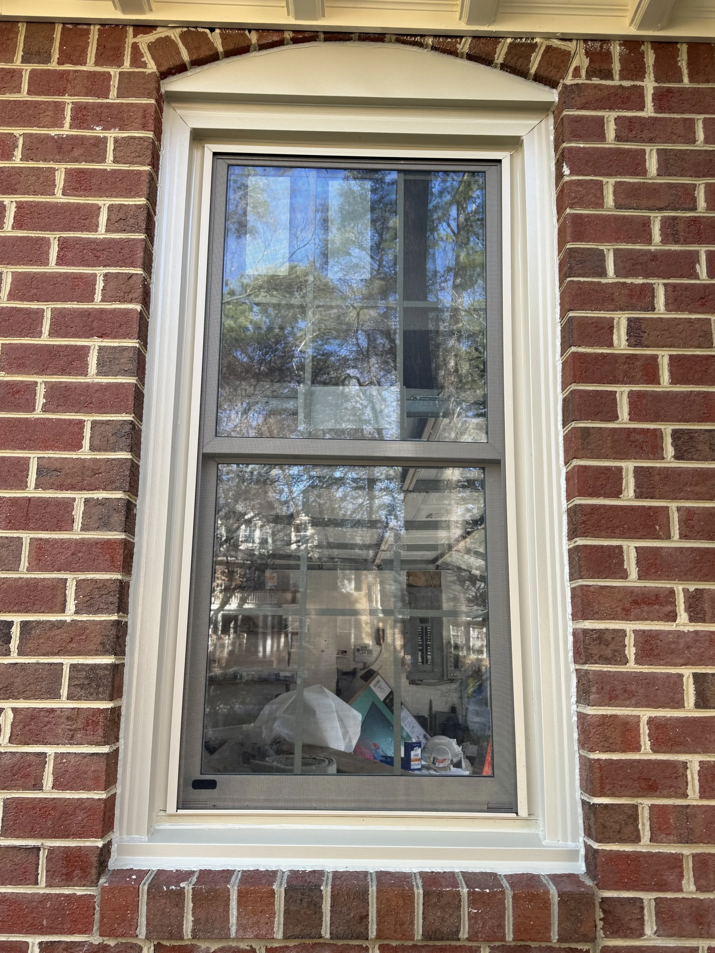 A brick house wall with a white-framed double-pane window that has a screen. The window reflects trees and the sky outside, with some miscellaneous items visible inside.