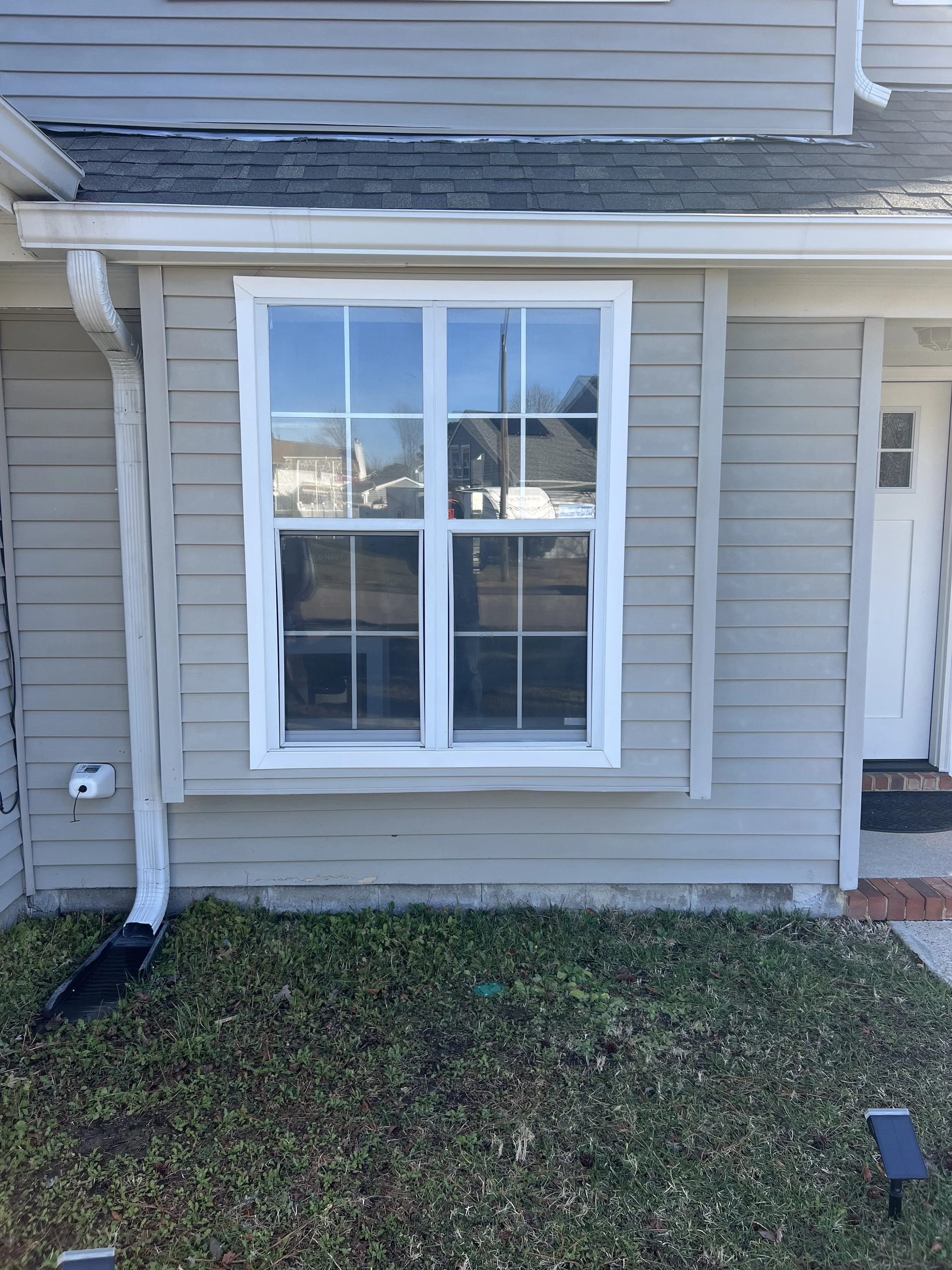 A beige house with vinyl siding and a large window with white framing, surrounded by green grass and some solar lights on the ground. A downspout is attached to the house and extends to a black gutter extension on the ground.