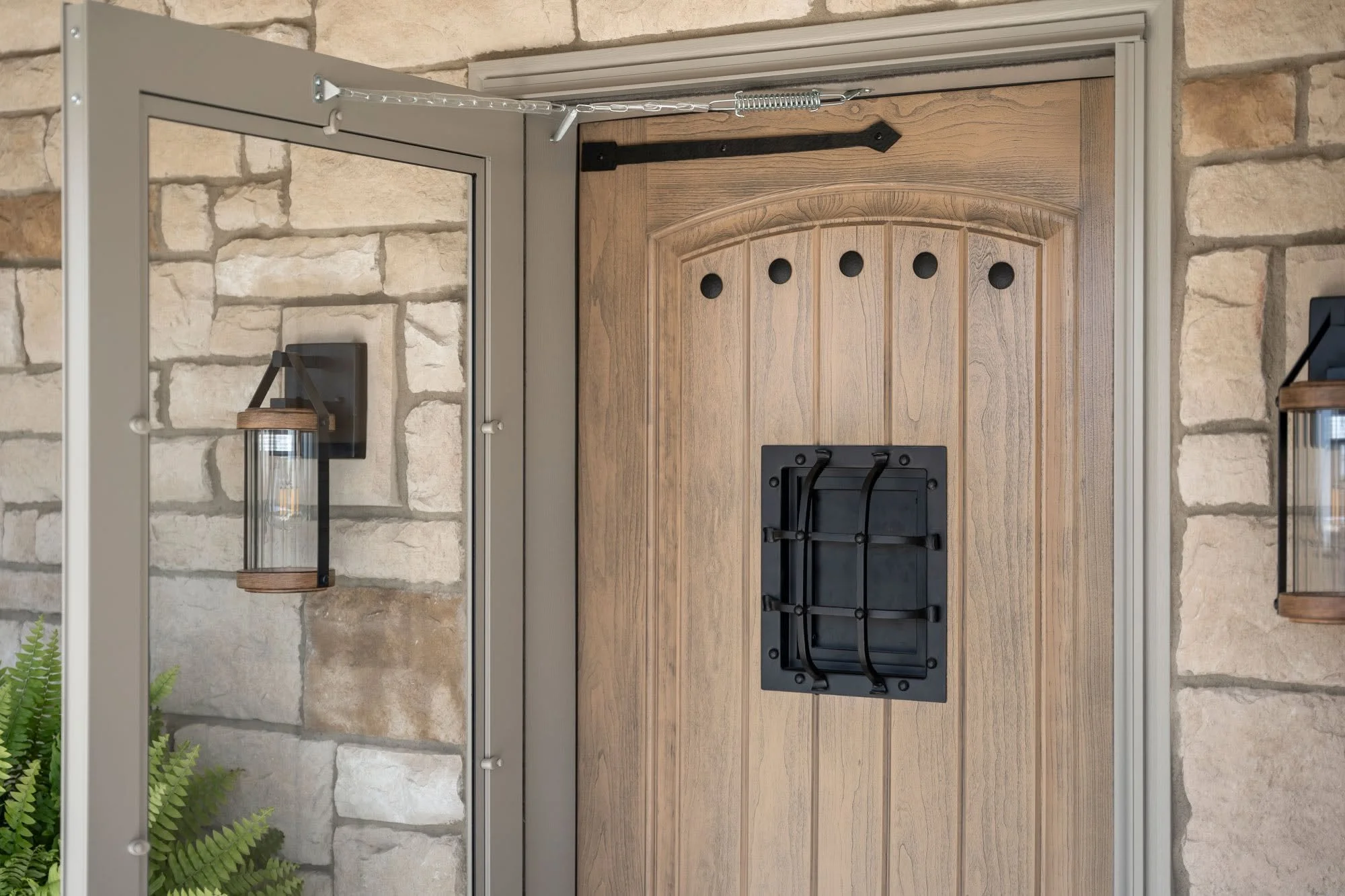 Exterior view of a front door with a wooden finish, black hardware, and a black mail slot, flanked by stone walls and a metal-framed mirror with lantern-style light fixtures on each side.