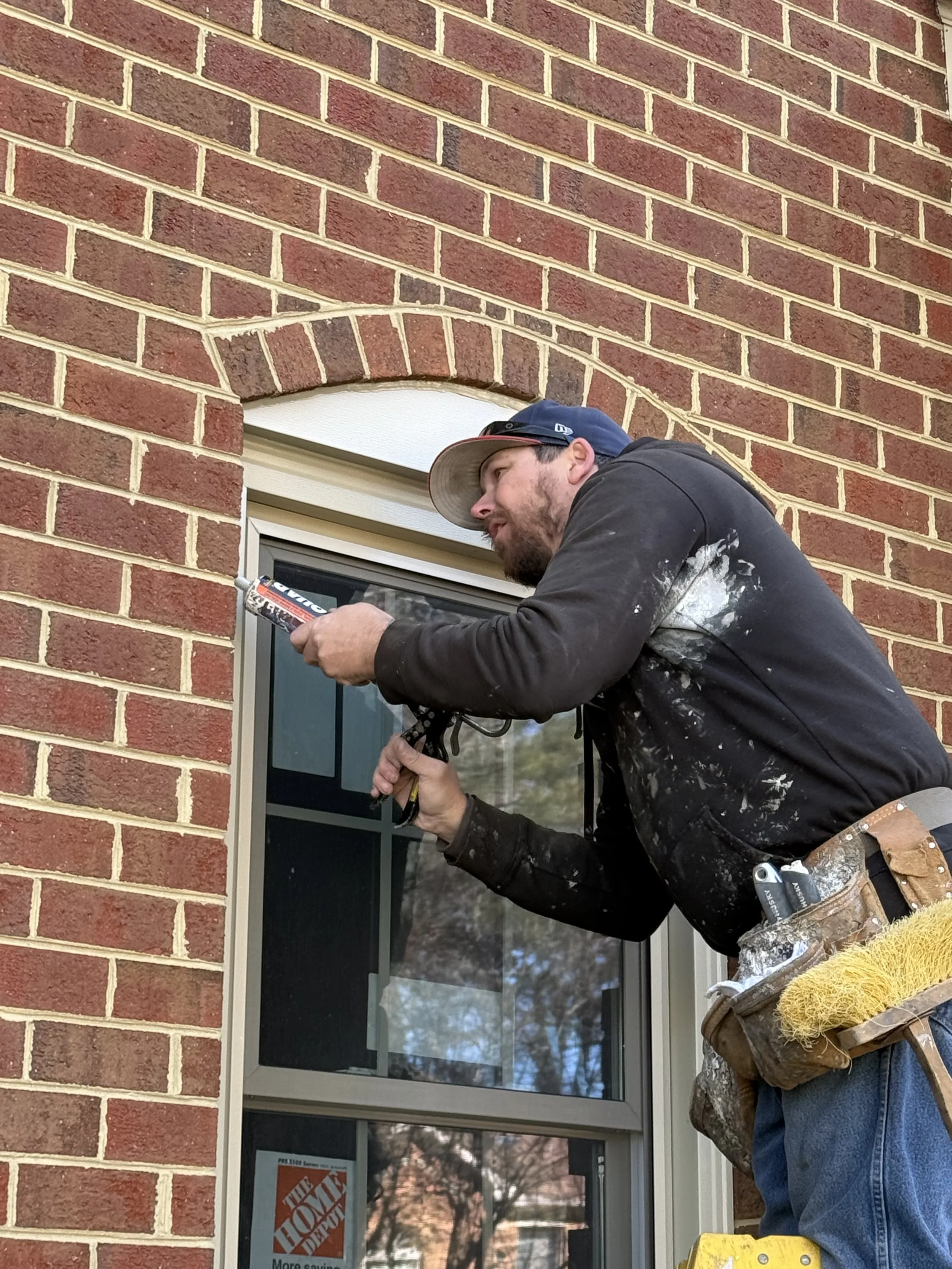 A man wearing a hat and work gloves is using caulk to seal the space between a brick wall and a window frame.