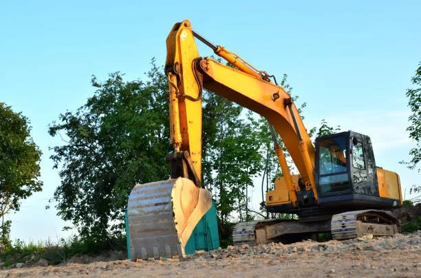 A yellow excavator with a heavy bucket attachment, parked on a dirt construction site with trees in the background.