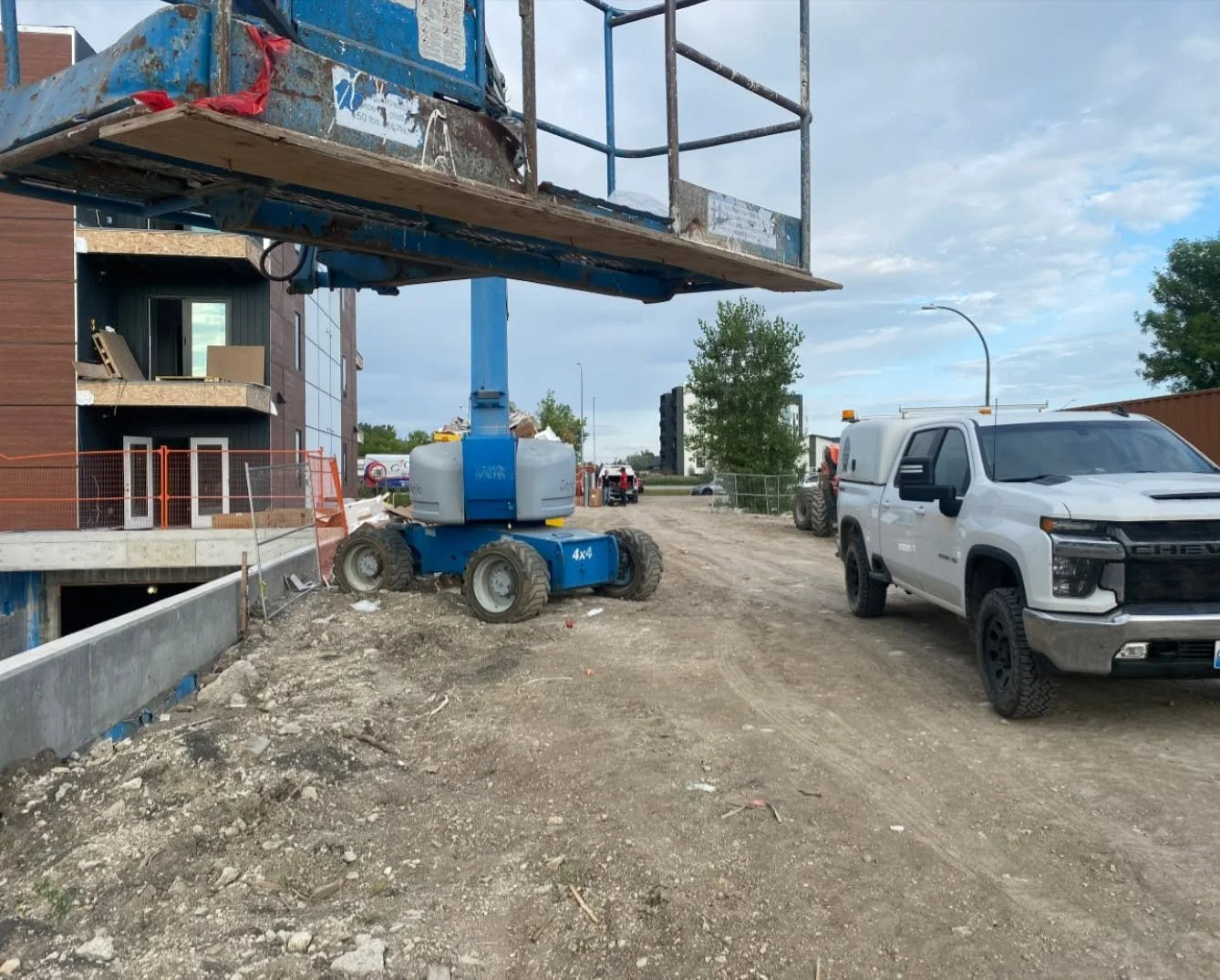 Construction site with a blue boom lift, a white pickup truck, and a modern building under construction. The ground is dirt and gravel, and there are construction materials and equipment scattered around.