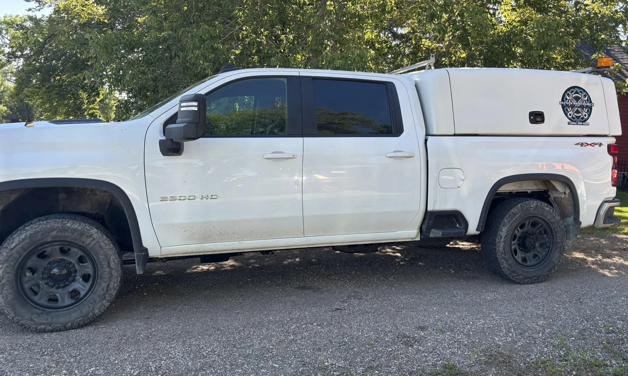 A white pickup truck with a logo on the side that reads 'Nex-Gen Maintenance,' parked on a dirt and gravel surface with green trees in the background.