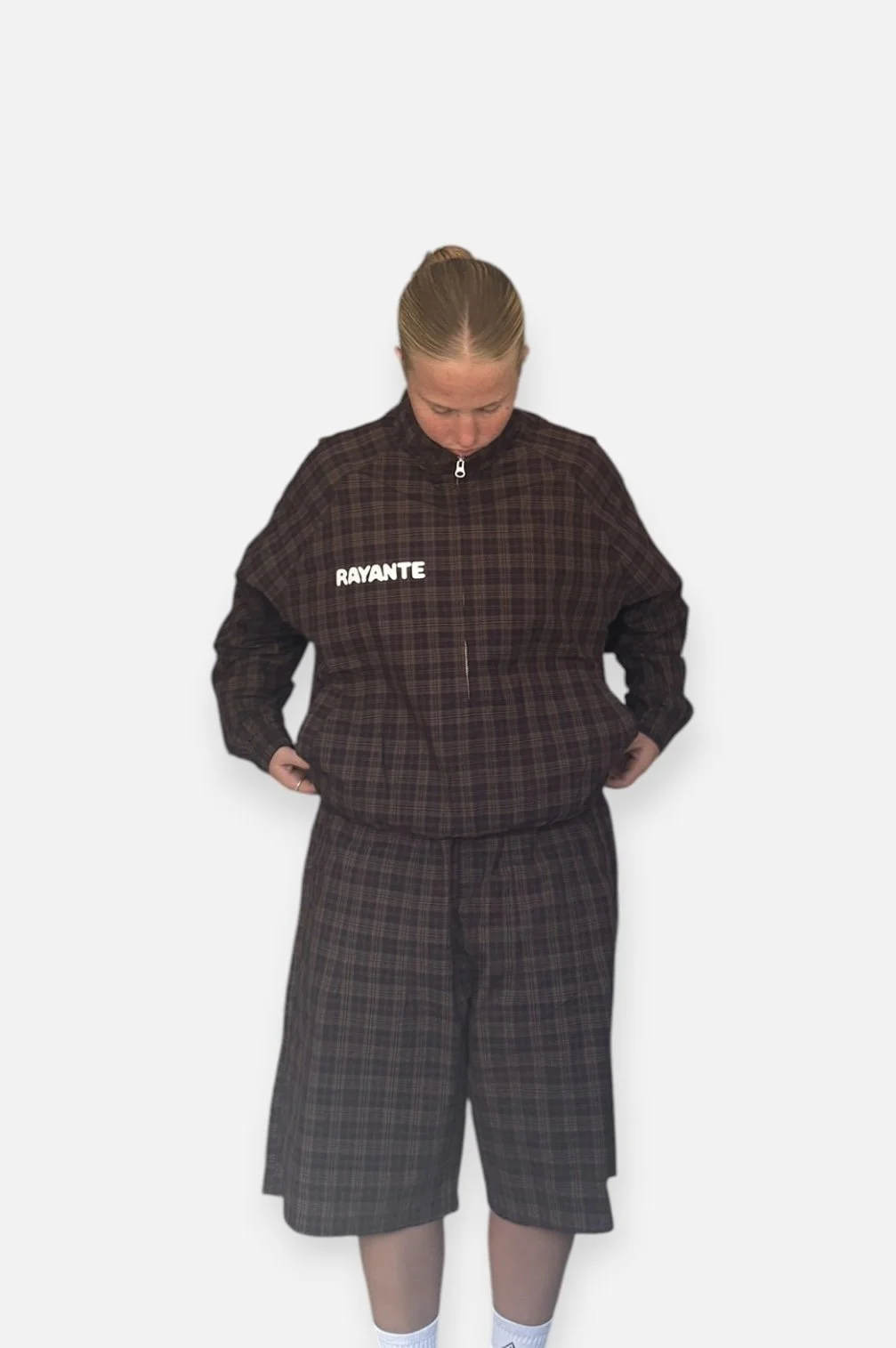 Woman wearing a dark plaid tracksuit with a jacket labeled 'RAYANTE', looking down with hands in pockets, standing against a plain white background.
