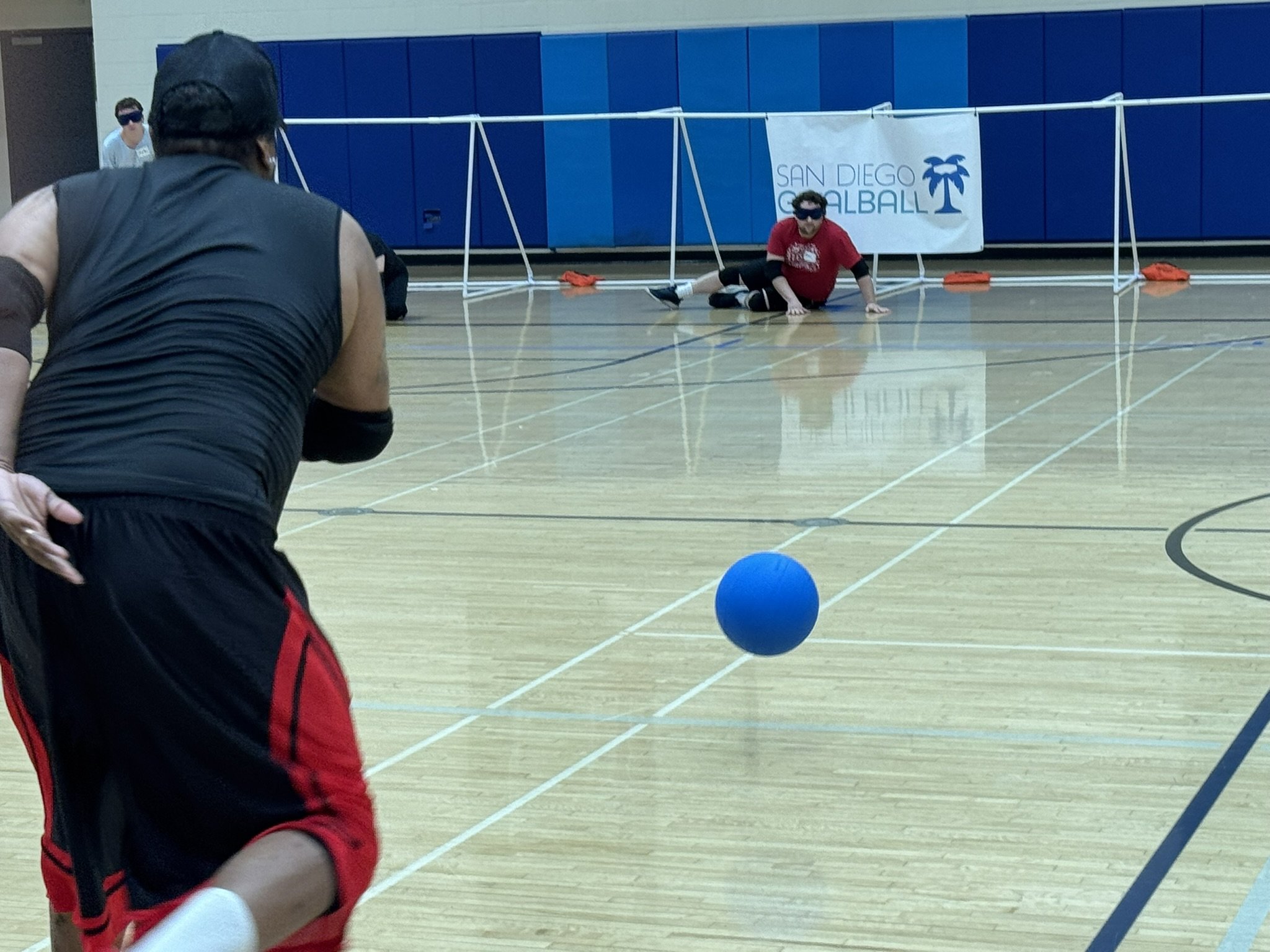 Antonio throwing a goalball towards an opposing player while trying to score a point. The San Diego Goalball banner is in the background.