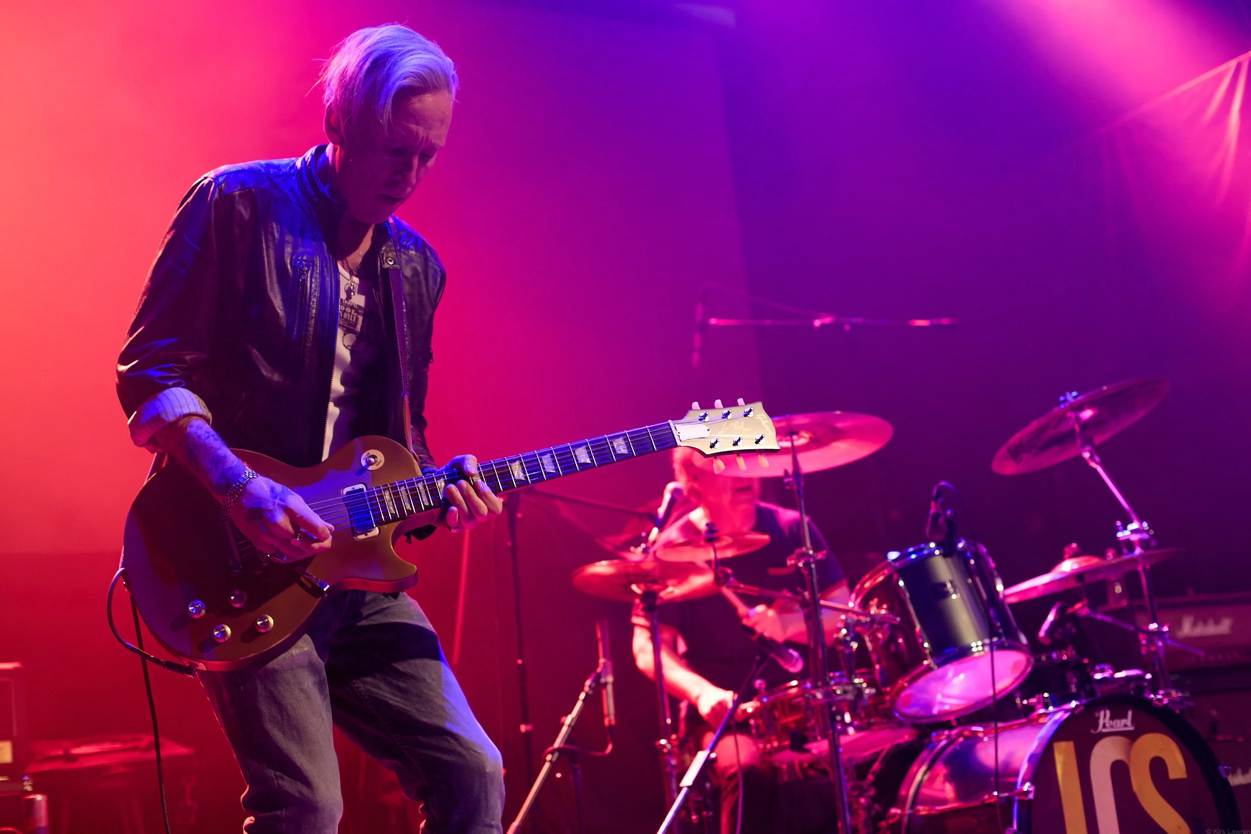 A musician playing an electric guitar on stage with colorful lighting, and a drummer in the background.