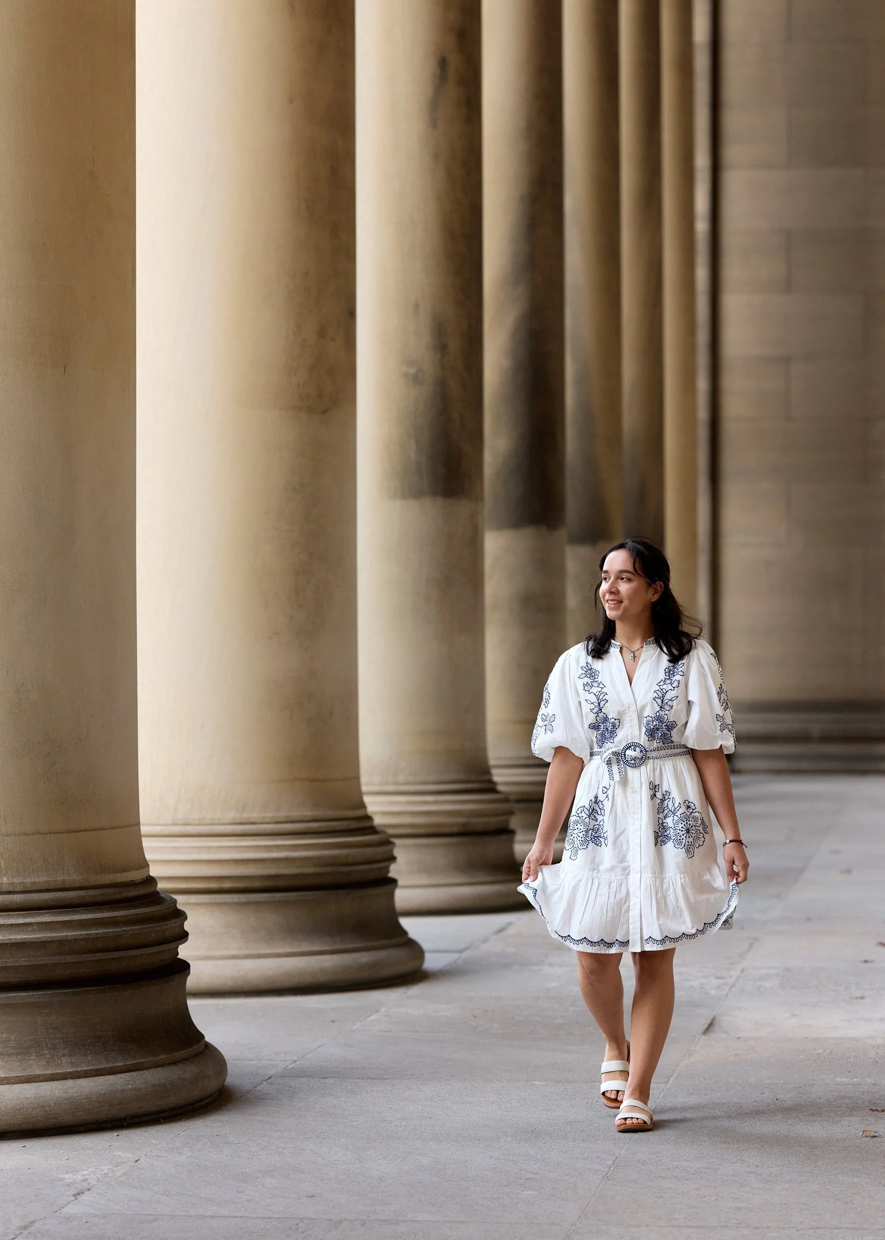 A young woman with dark hair wearing a white dress with blue embroidery walking along a colonnade with large stone columns.
