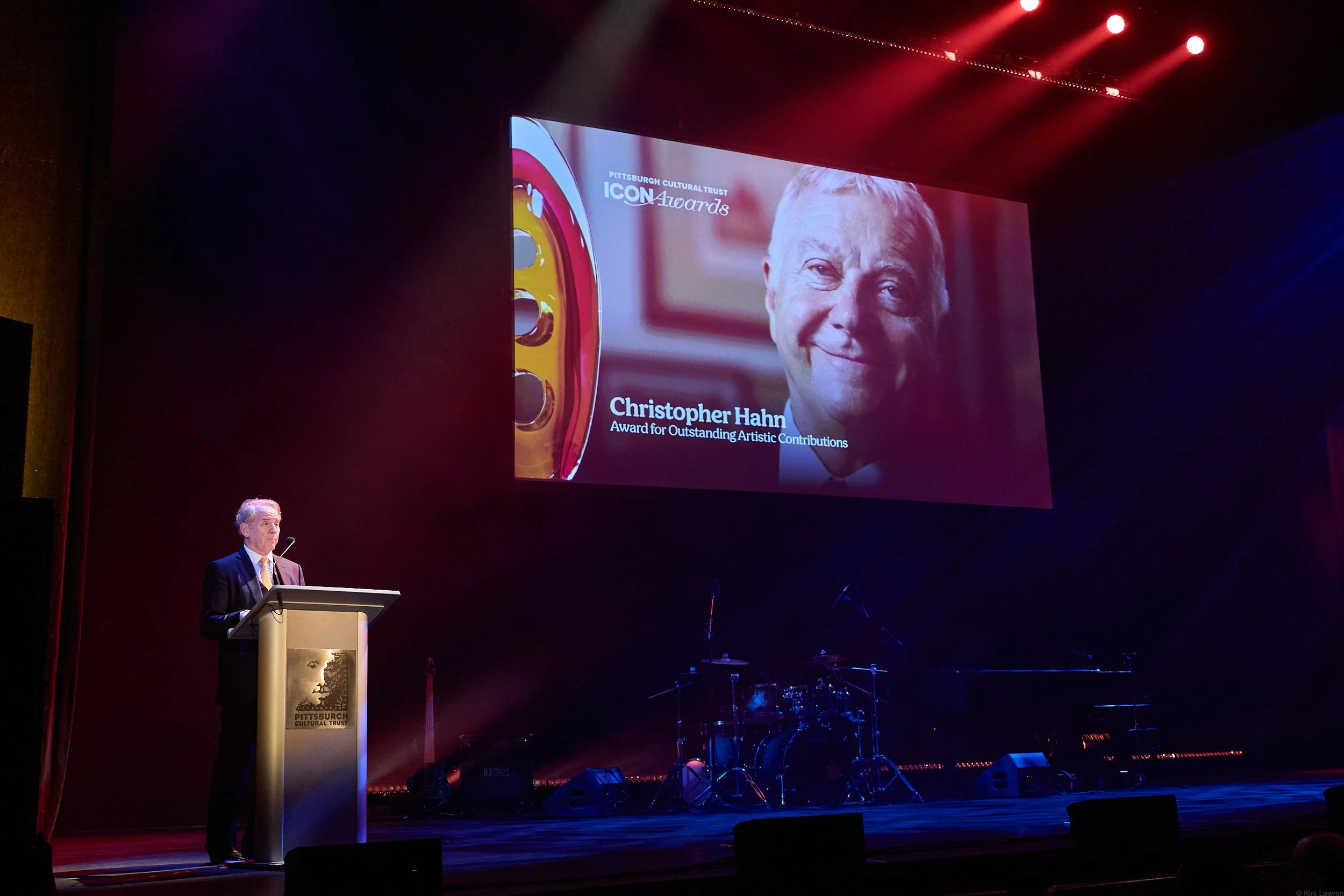 A man in a suit standing at a podium on a stage during an awards ceremony. A large screen behind him displays a close-up photo of a smiling man with text identifying him as Christopher Hahn receiving the 'Award for Outstanding Artistic Contributions'
