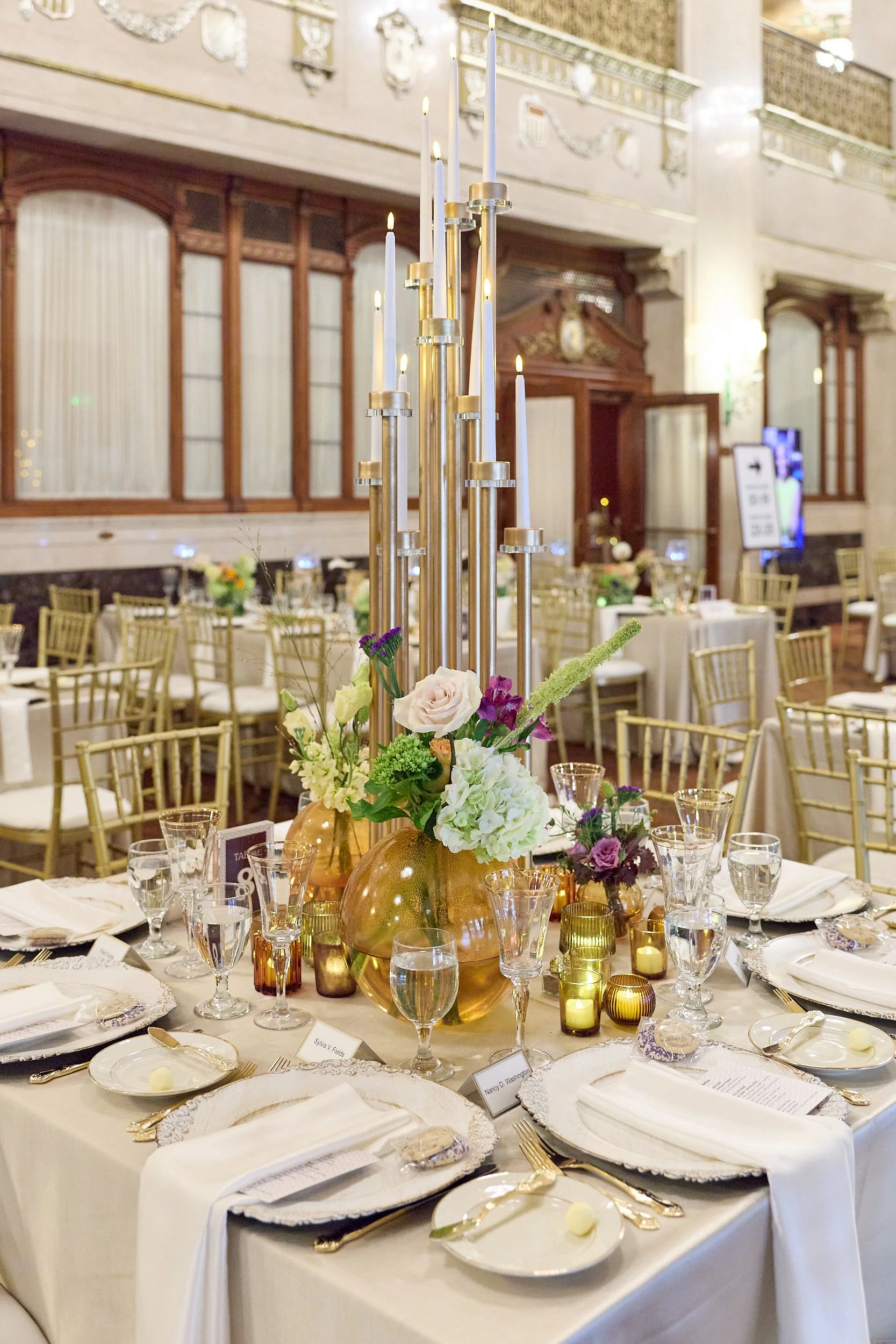 Elegant banquet table with floral centerpiece, golden accents, candlesticks, and place settings in a grand hall.