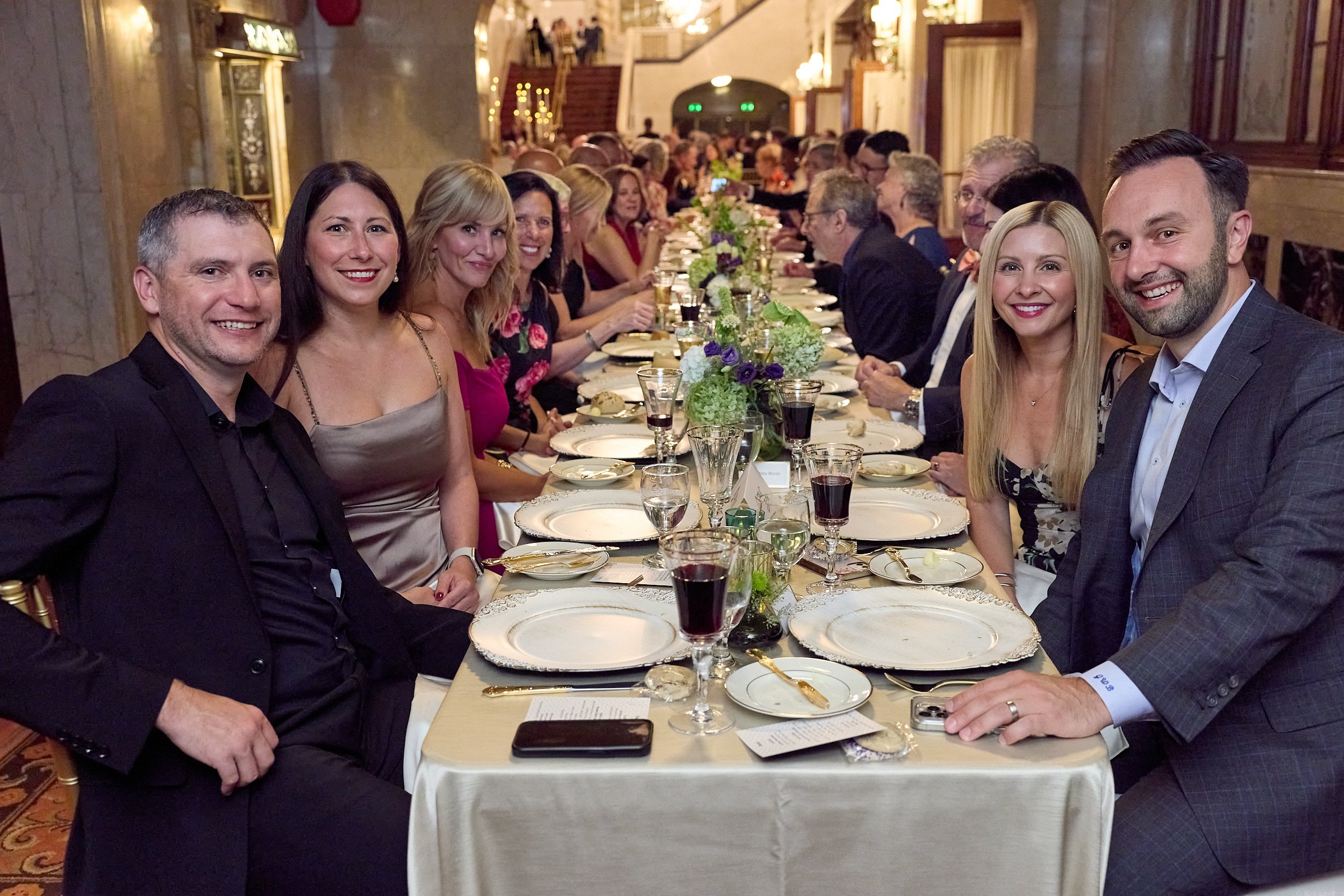 People sitting around a dinner table at a formal event, smiling for the camera, with elegant table settings and floral centerpieces in a decorated banquet hall.
