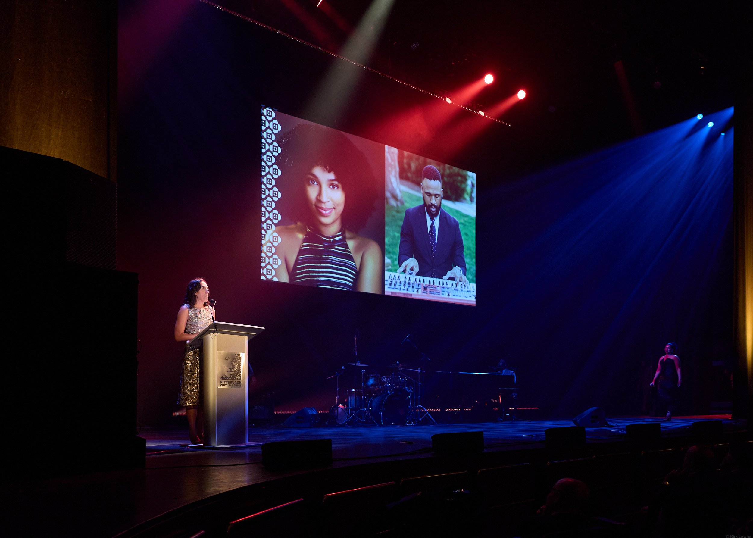A woman is speaking at a podium on a stage with a large screen behind her displaying two photographs: one of a woman with curly hair and a striped top, and one of a man with short hair and a suit playing a keyboard. The stage is dimly lit with blue a