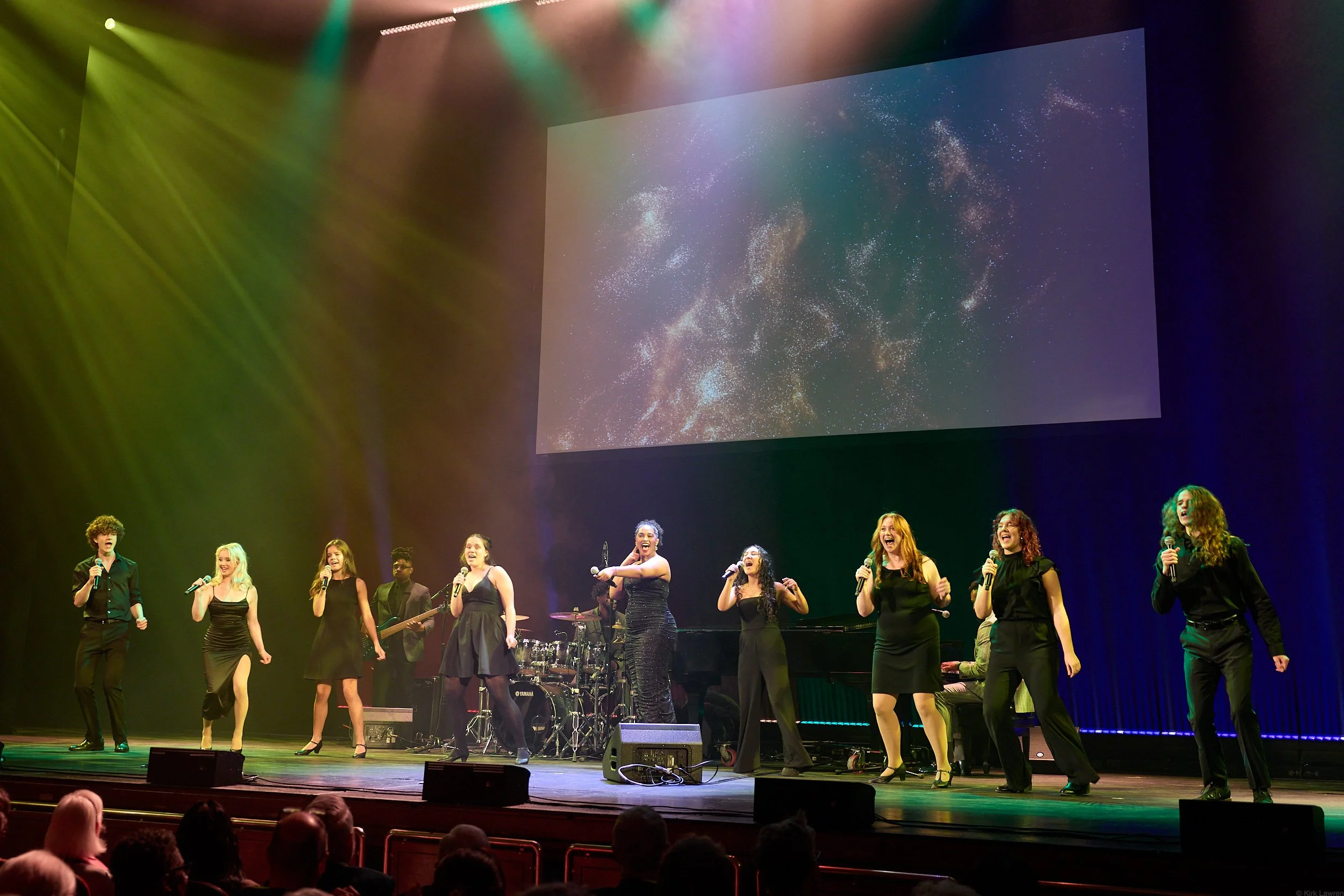 A group of diverse singers on stage performing with microphones, a drummer in the background, and a backdrop displaying a starry galaxy.